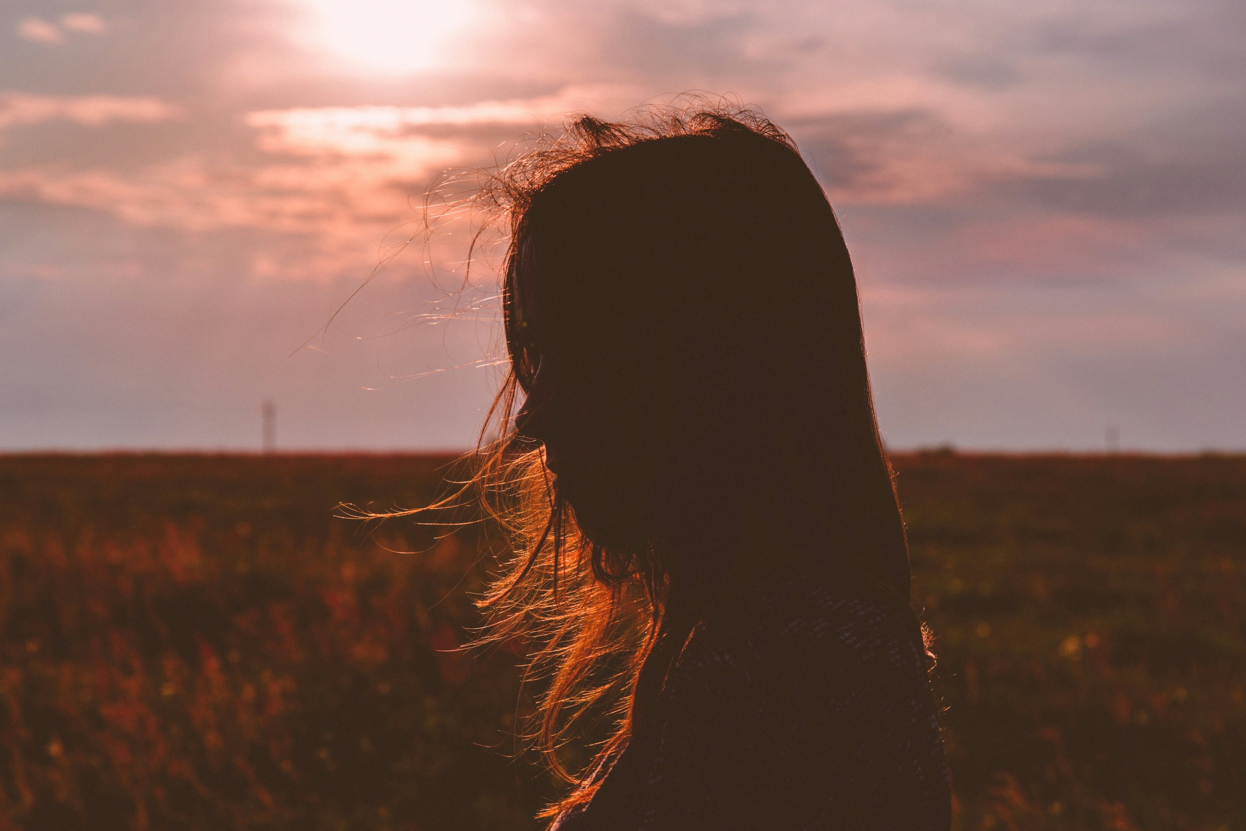 Silhouette of a woman with long hair standing in a field during sunset, with clouds and a sunset sky in the background.
