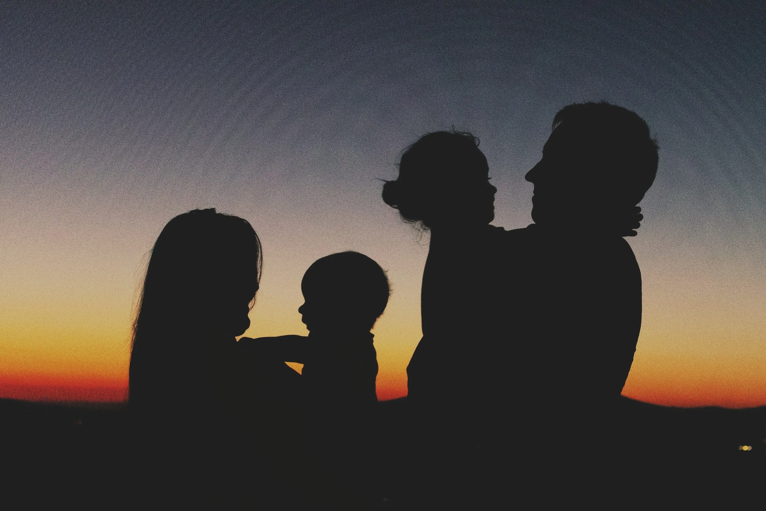 Silhouettes of a family with two children against a sunset sky, with one parent holding a baby, outdoor scene