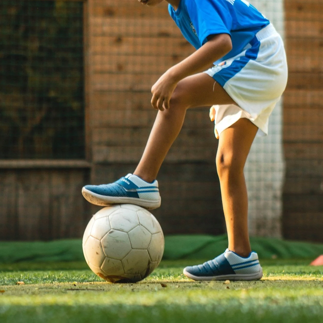 A young person balancing a soccer ball with their foot on a field, wearing blue sports shoes, blue shirt, and white shorts.