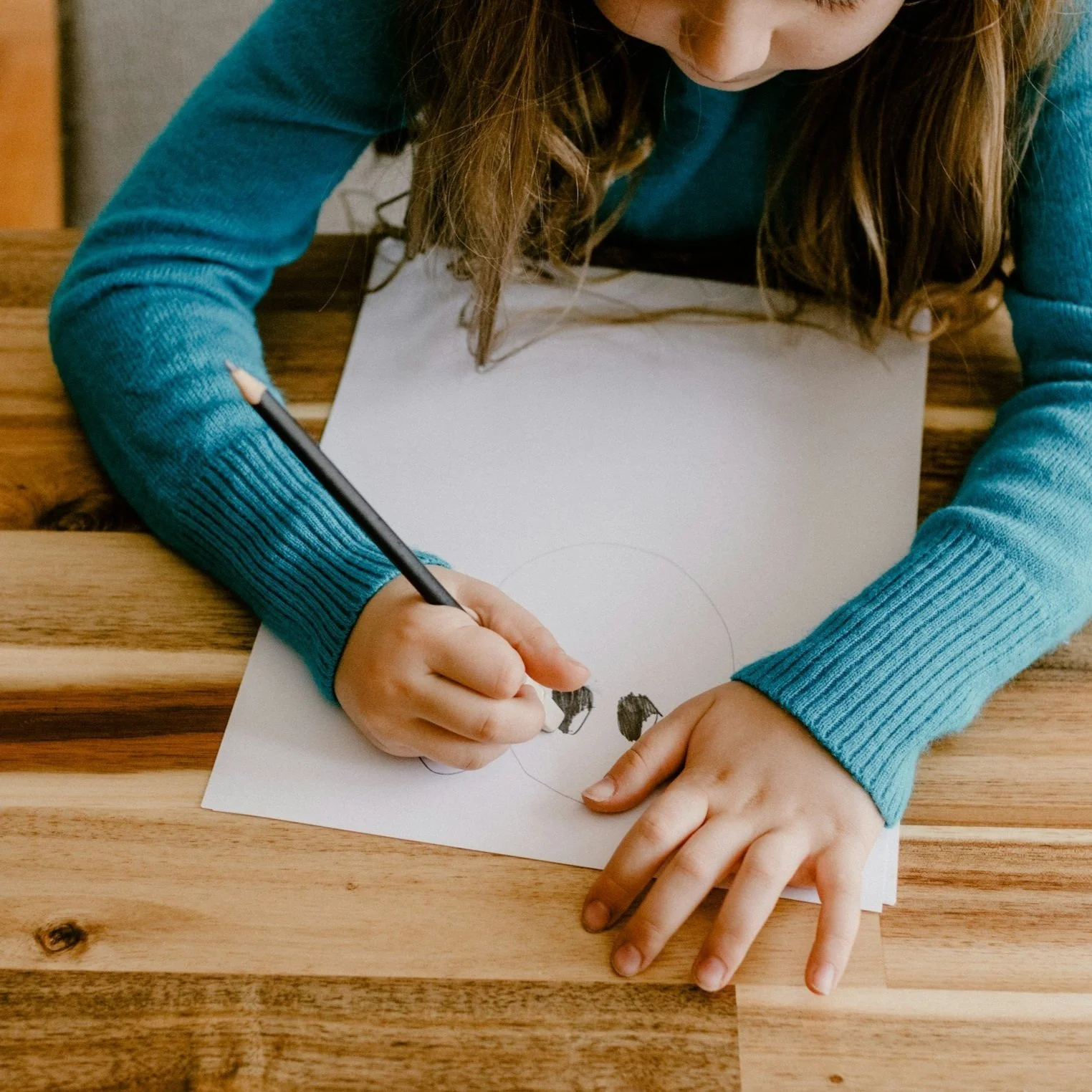 A young girl in a blue sweater drawing a panda face on a white sheet of paper at a wooden table.