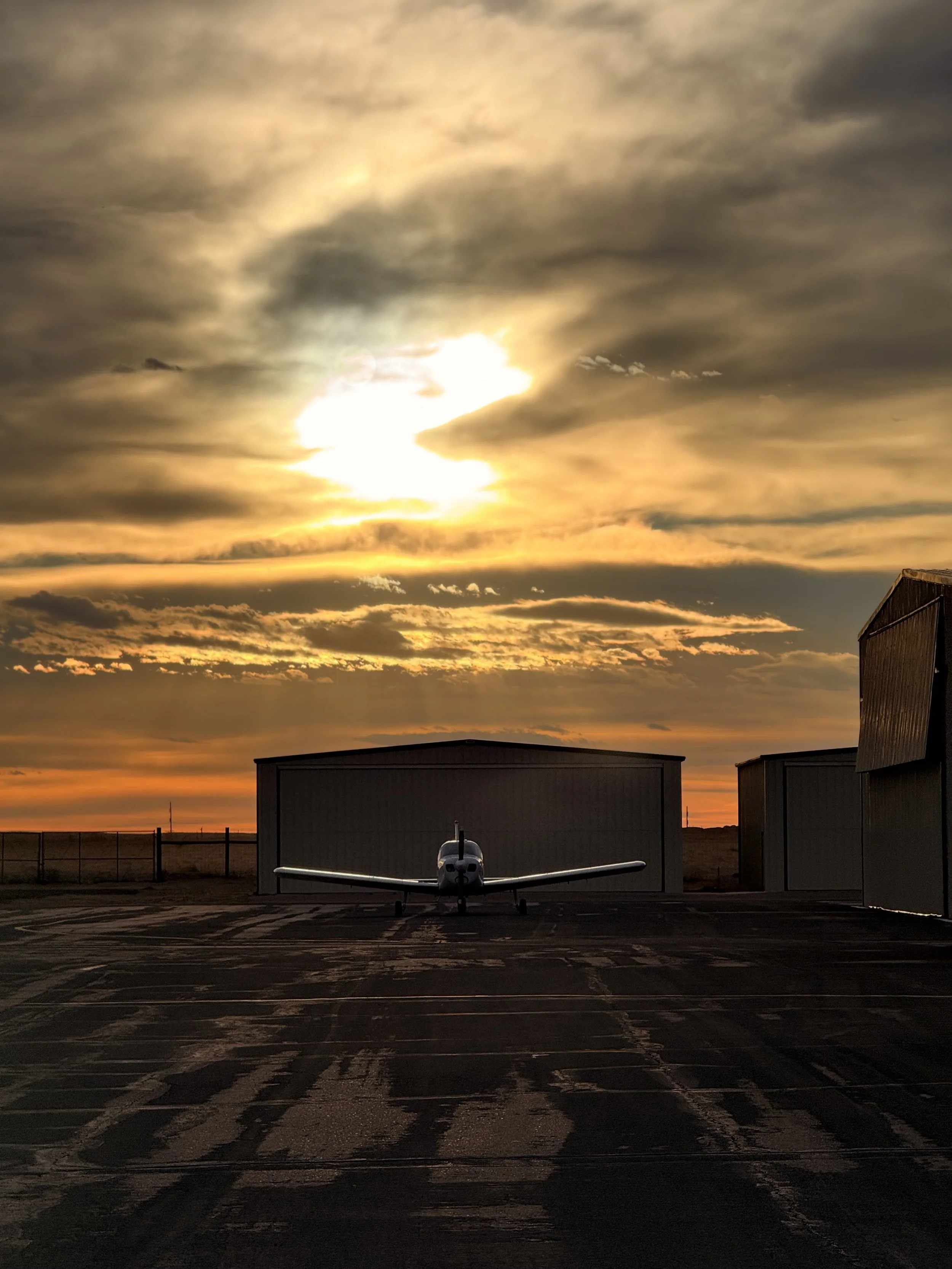 Small airplane parked in front of a hangar during a dramatic sunset, with vibrant clouds and golden sky.