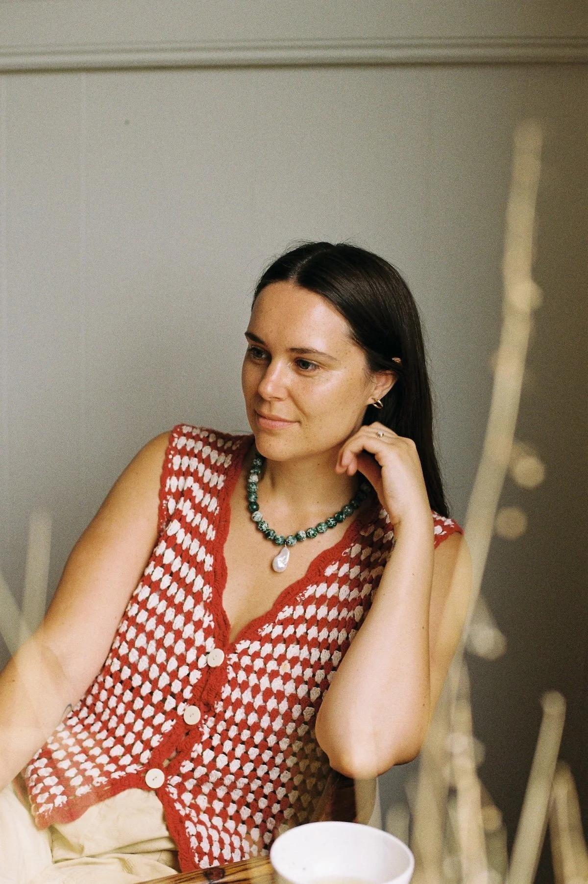 Person wearing a red and white crochet vest with a green beaded necklace, sitting at a table with a cup.