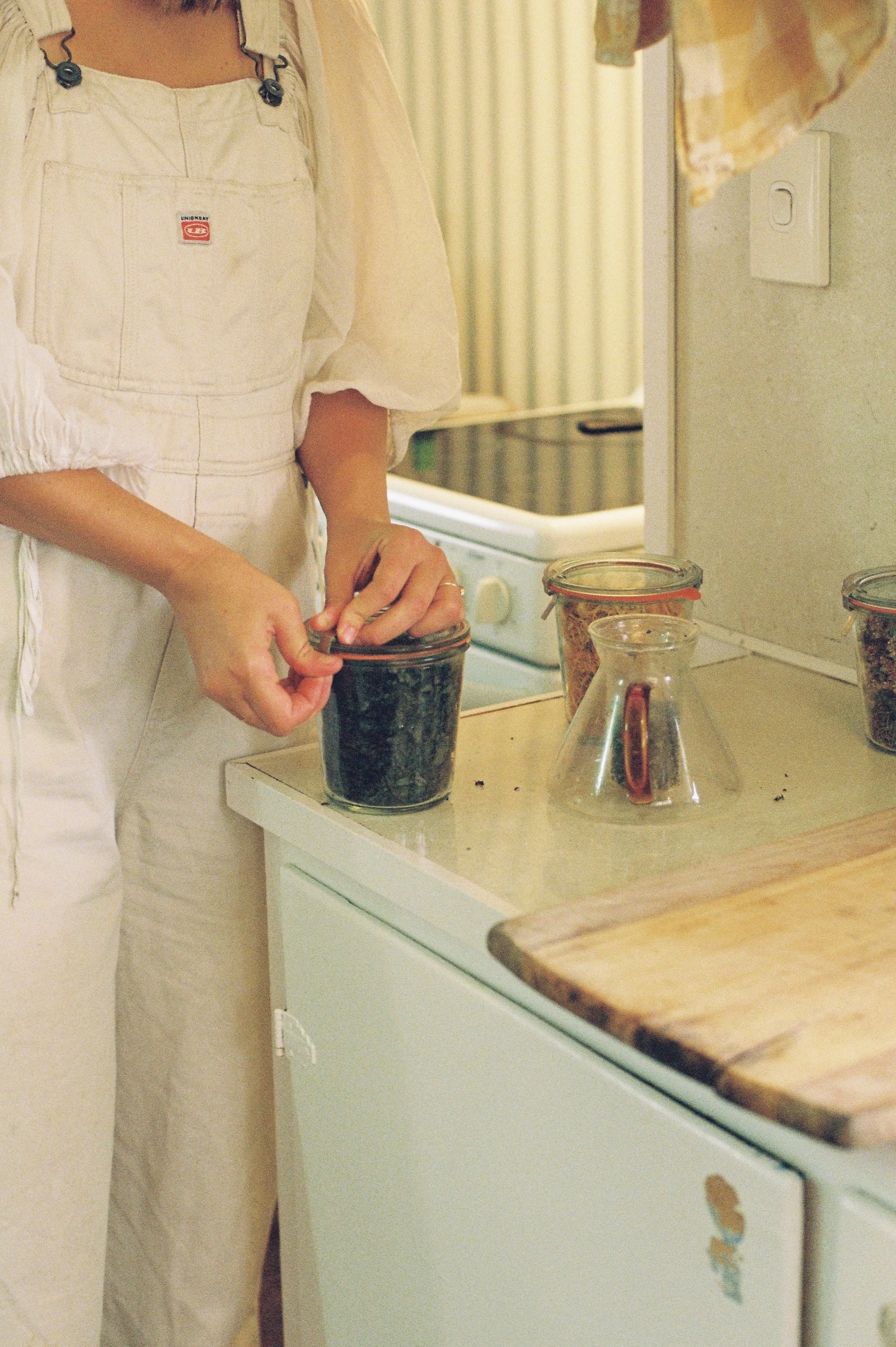 Person in white apron opening glass jar on kitchen counter with cutting board and jars nearby.