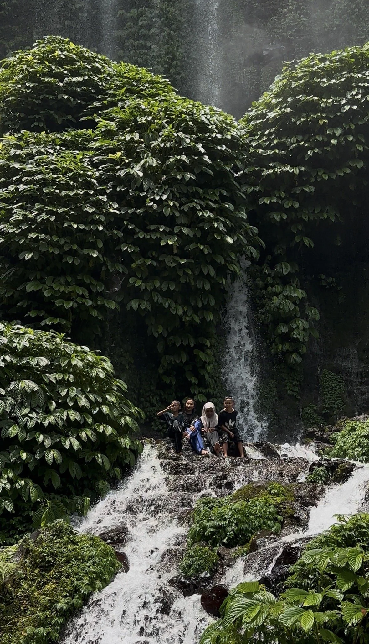 Local children sitting beside a tropical waterfall in Lombok surrounded by lush jungle scenery and flowing freshwater streams.