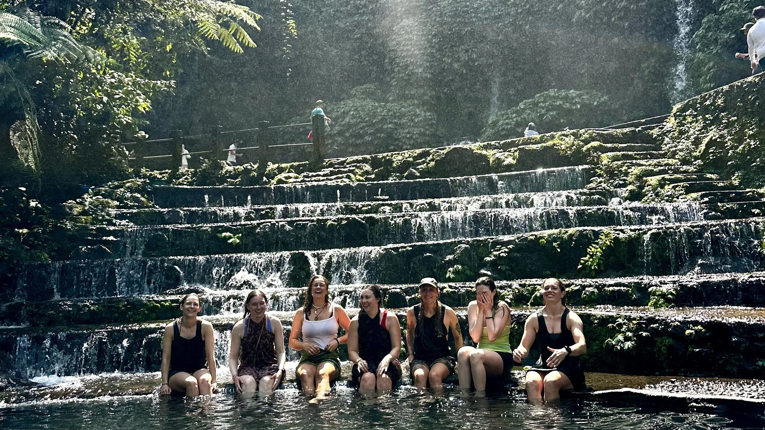 Group of female travellers relaxing beneath the cascading Benang Kelambu Waterfall in Lombok surrounded by lush tropical jungle scenery.