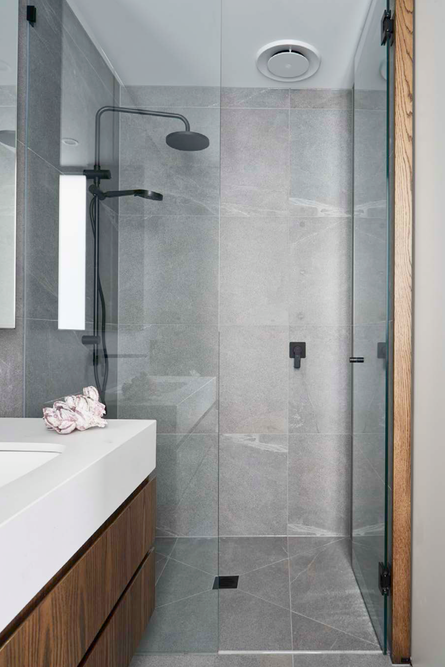 Modern bathroom with gray tiled shower area, black shower fixtures, wooden storage cabinet, and white countertop with pink fabric swatch, viewed through a glass shower door.