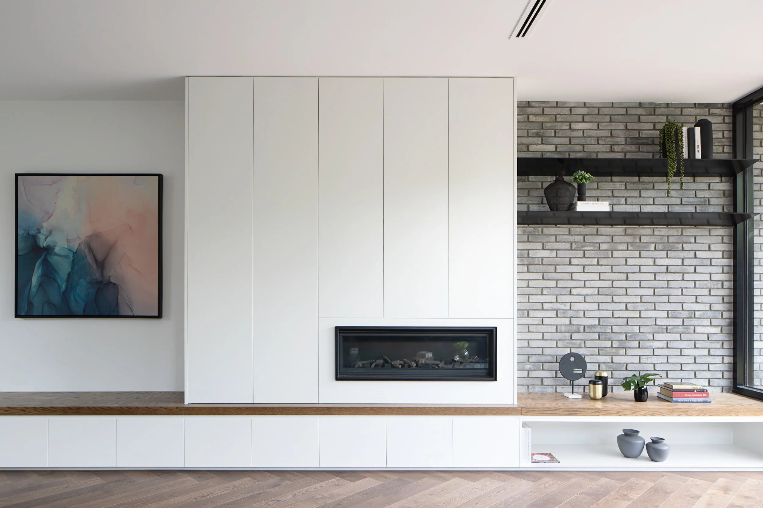 Modern living room with a white wall, abstract art, brick fireplace, black shelving holding plants and books, and decorative objects on a wood floor.