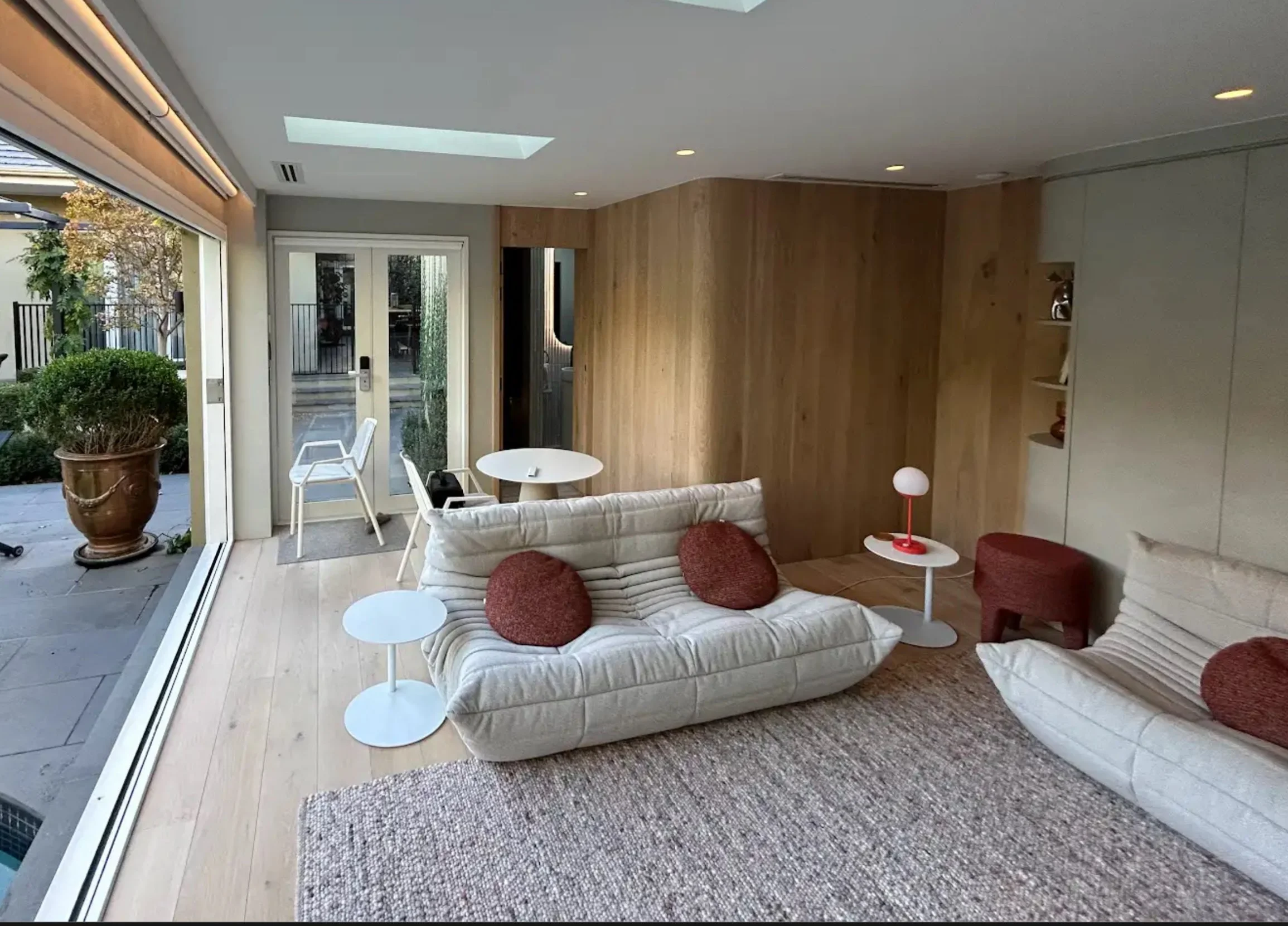 Living room with large glass sliding door leading to outdoor patio, beige sofa with red pillows, small white tables, area rug, wood-paneled wall, and a skylight in the ceiling.