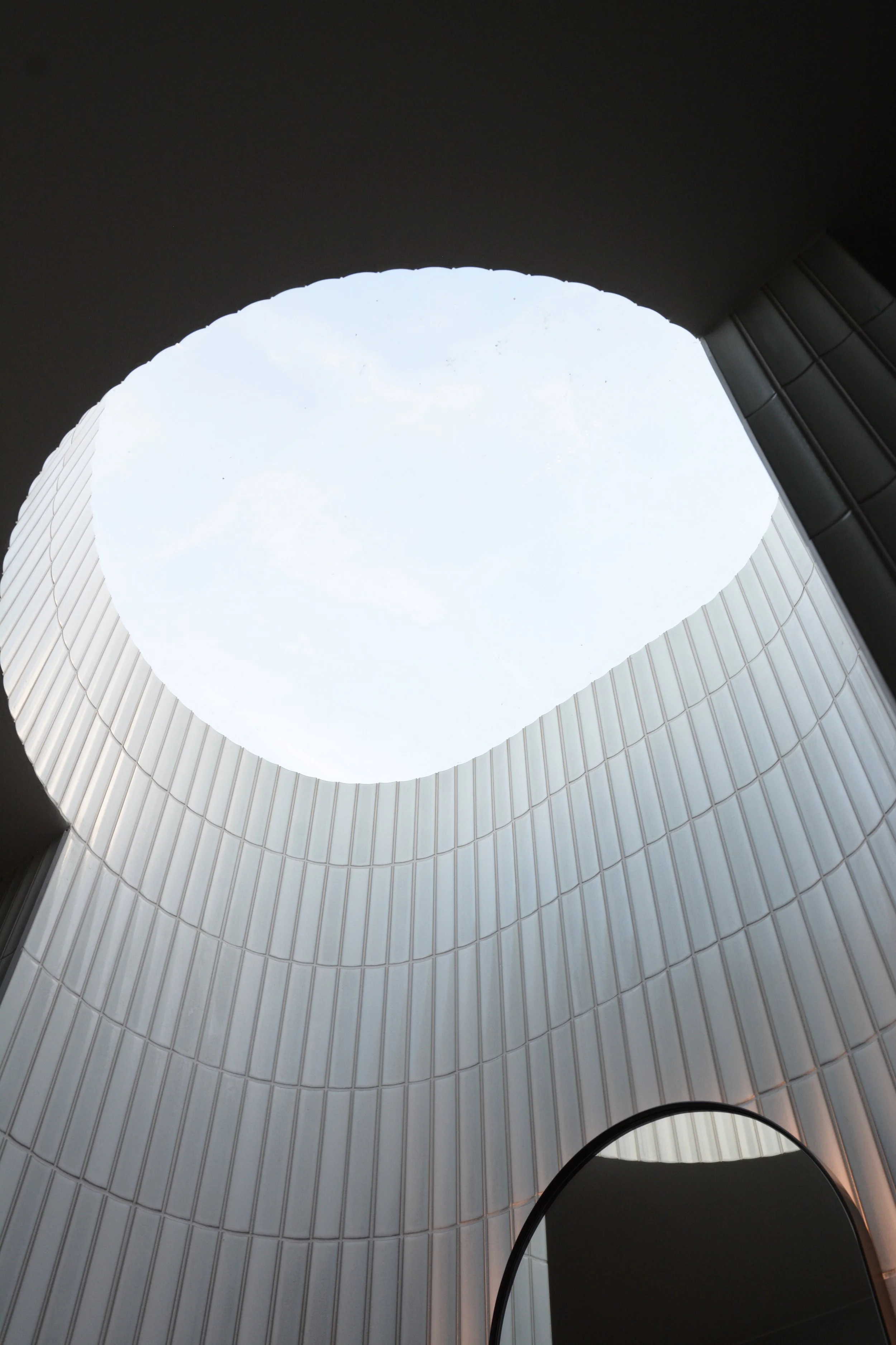 Looking up at the sky from inside a tall, cylindrical building with white tiled walls, with a circular opening at the top showing a cloudy sky.