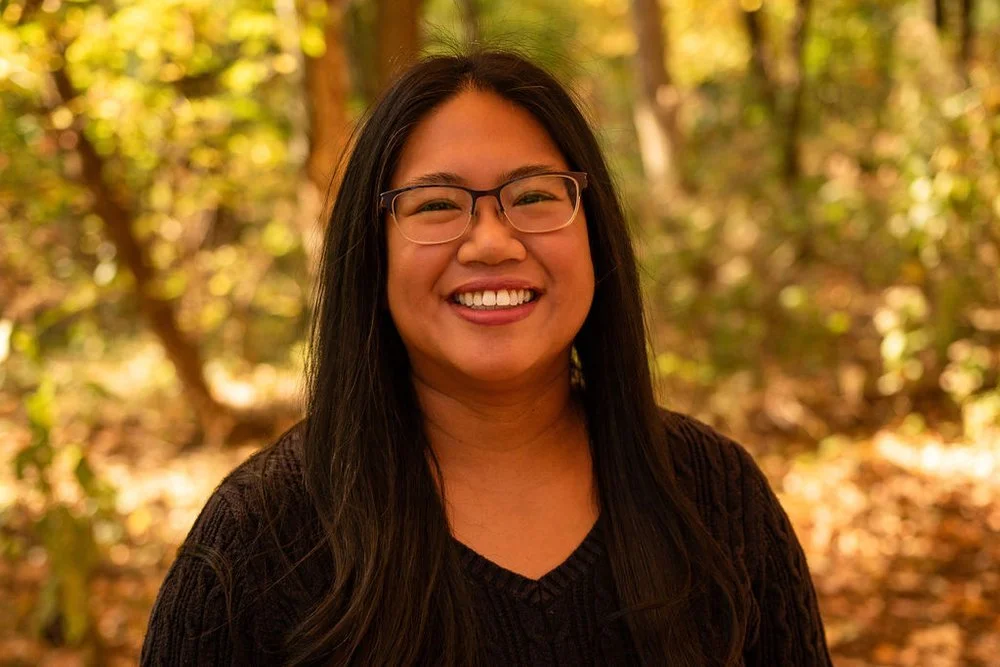 A cheerful Asian woman with long dark brown hair smiling outdoors in a forest during autumn with fall foliage in the background.
