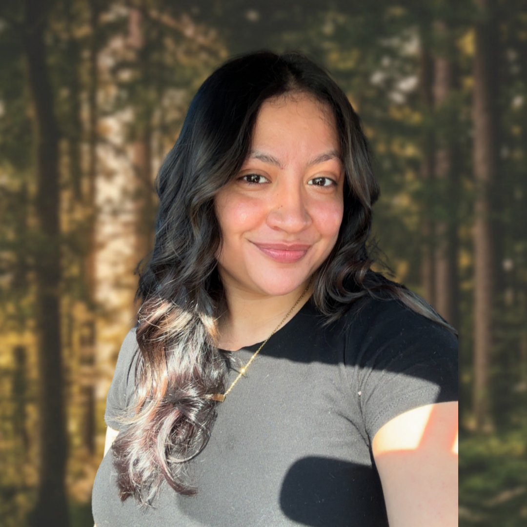 A woman with long, dark, wavy hair smiling, wearing a black top and gold necklace, with a blurred outdoor forest background.