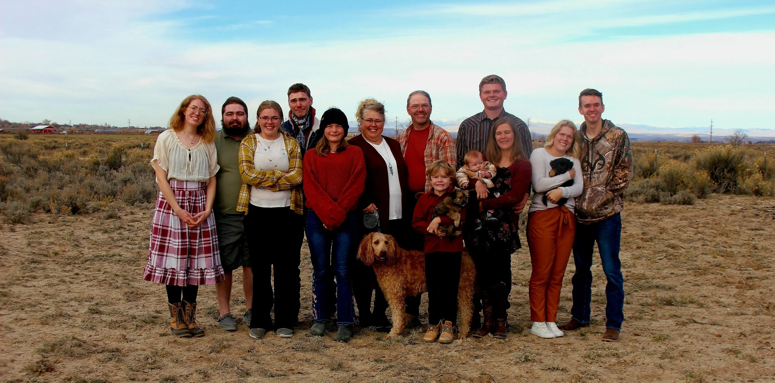 A large group of people and dogs standing outdoors in a field with mountains and cloudy sky in the background.