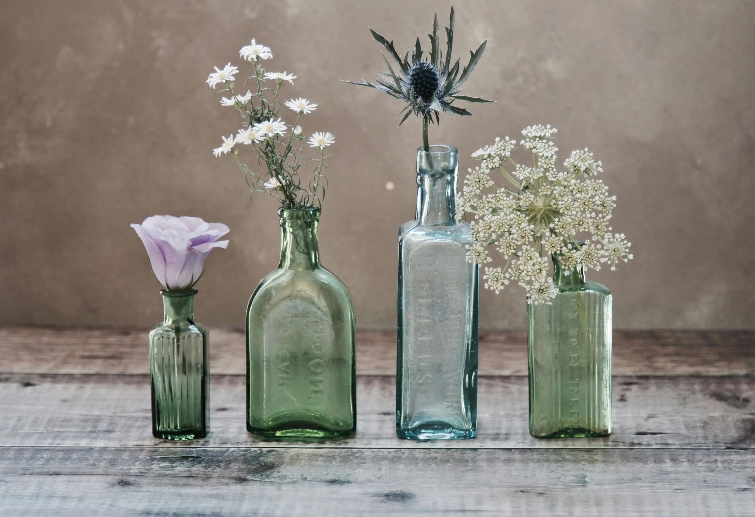Four vintage glass bottles with different small flowers on a rustic wooden surface.