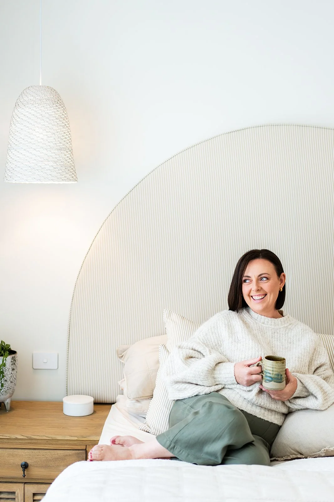 A woman sitting in bed, holding a coffee mug, smiling, in a cozy bedroom with a padded headboard and a bedside table with a plant and a small white object.