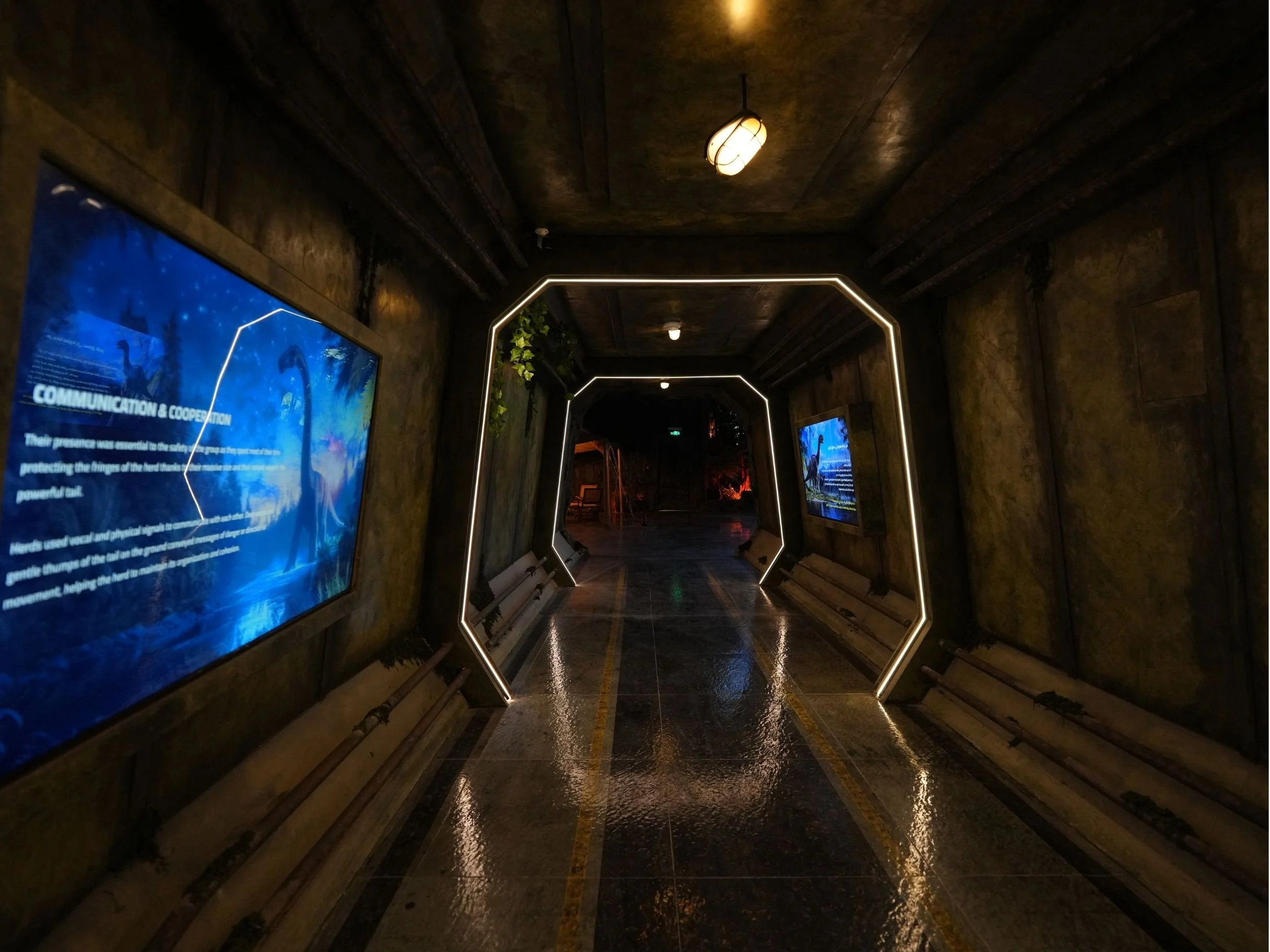 Underground tunnel with digital screens on the left side, illuminated geometric arch, wet floor reflecting lights, and dark, reinforced concrete walls and ceiling.