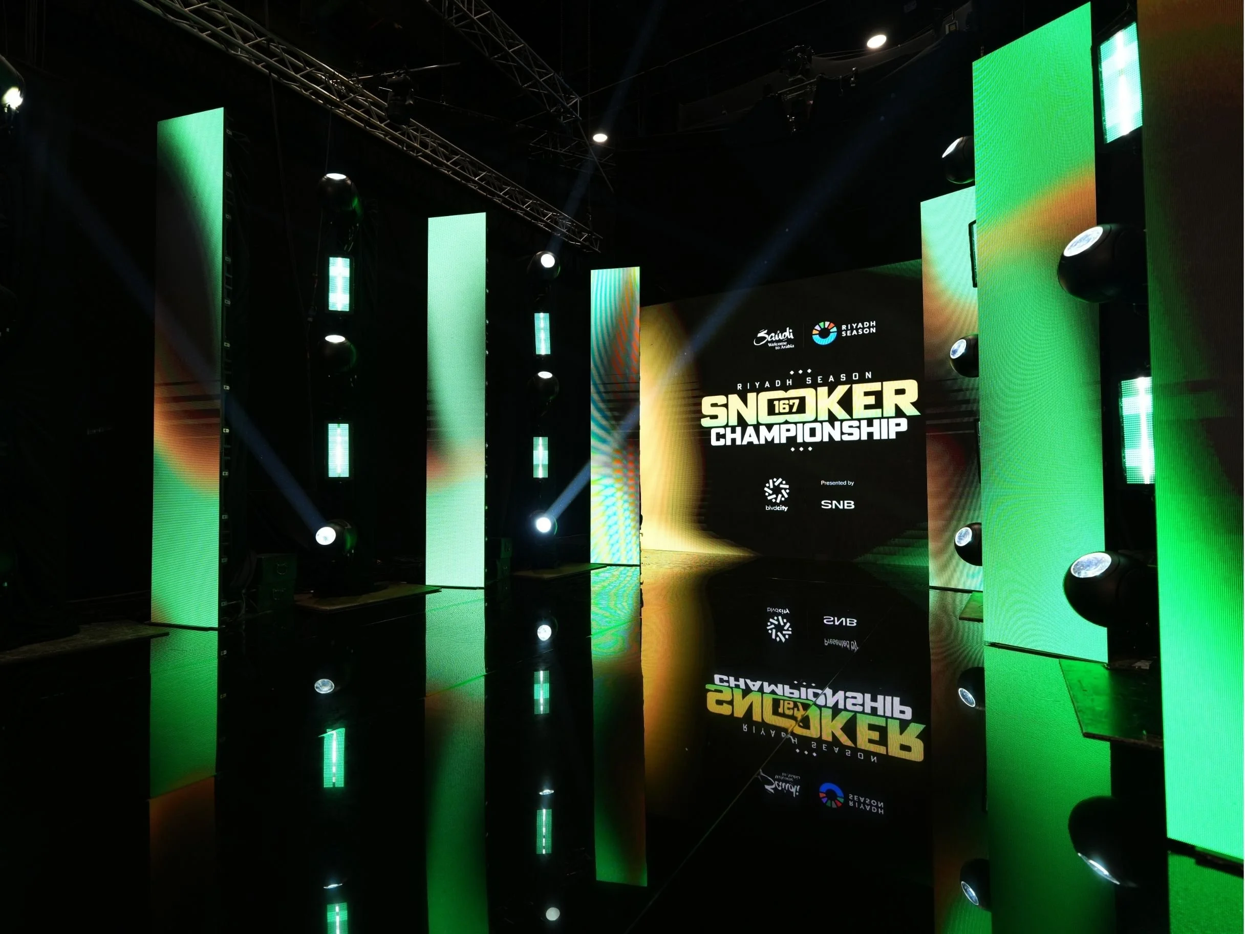 Stage with multiple large vertical digital screens displaying green patterns and event branding for Riyadh Season Snooker Championship. The stage is equipped with stage lights and a large digital display showing event logos, with reflections visible 