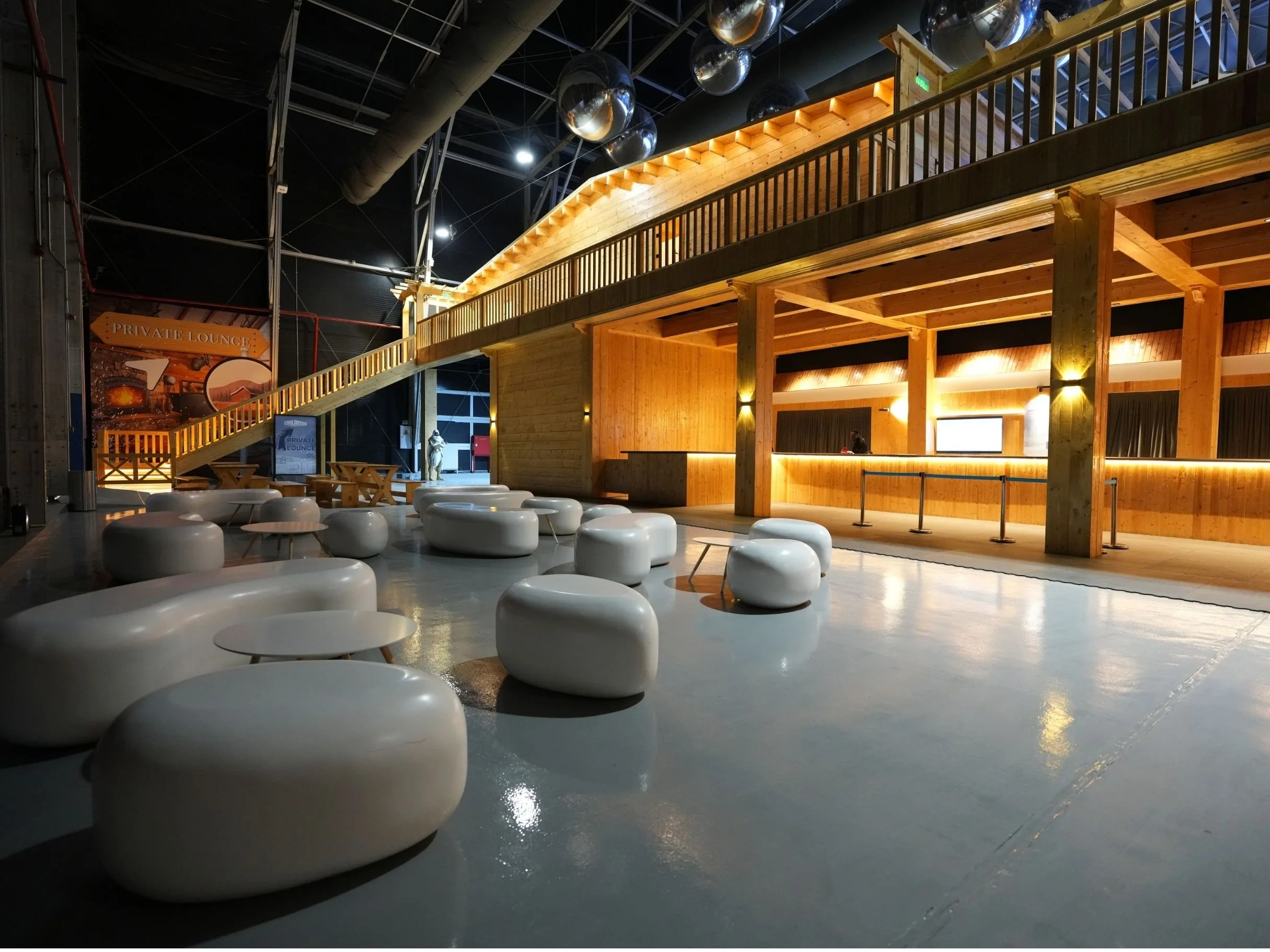 Interior of a modern building with wooden accents, a mezzanine, and a reception counter. There are white, rounded seating pods on a smooth floor. The ceiling features large metallic spheres and exposed ducts. cool arena iceskating rink.