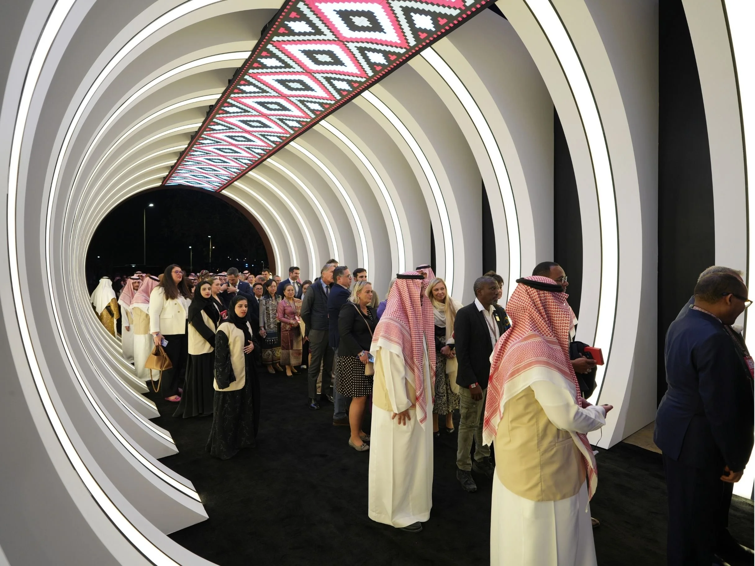 A diverse group of people, including Middle Eastern and Western individuals, in traditional and formal attire, gathered inside a modern, well-lit tunnel with glowing white arches and a patterned ceiling at night.boulevard world, sela, construction