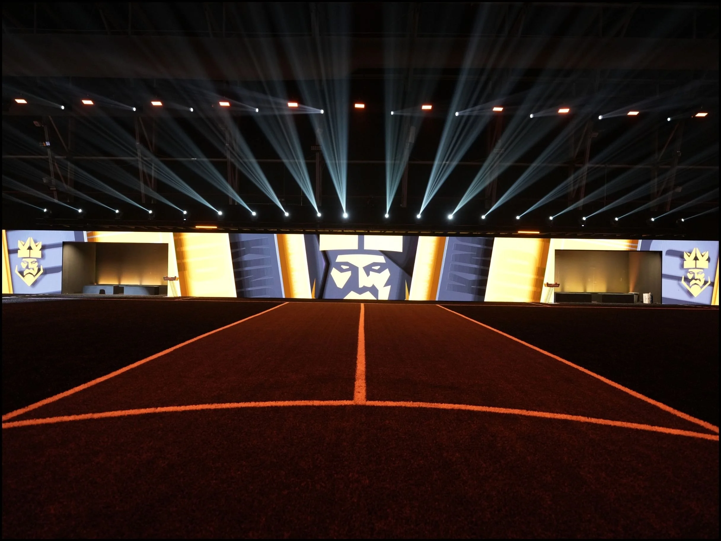 An indoor stage with bright lights and large screens featuring a logo with a face and crown, framed by decorative banners, and a section of red carpet with orange lines leading toward the stage.