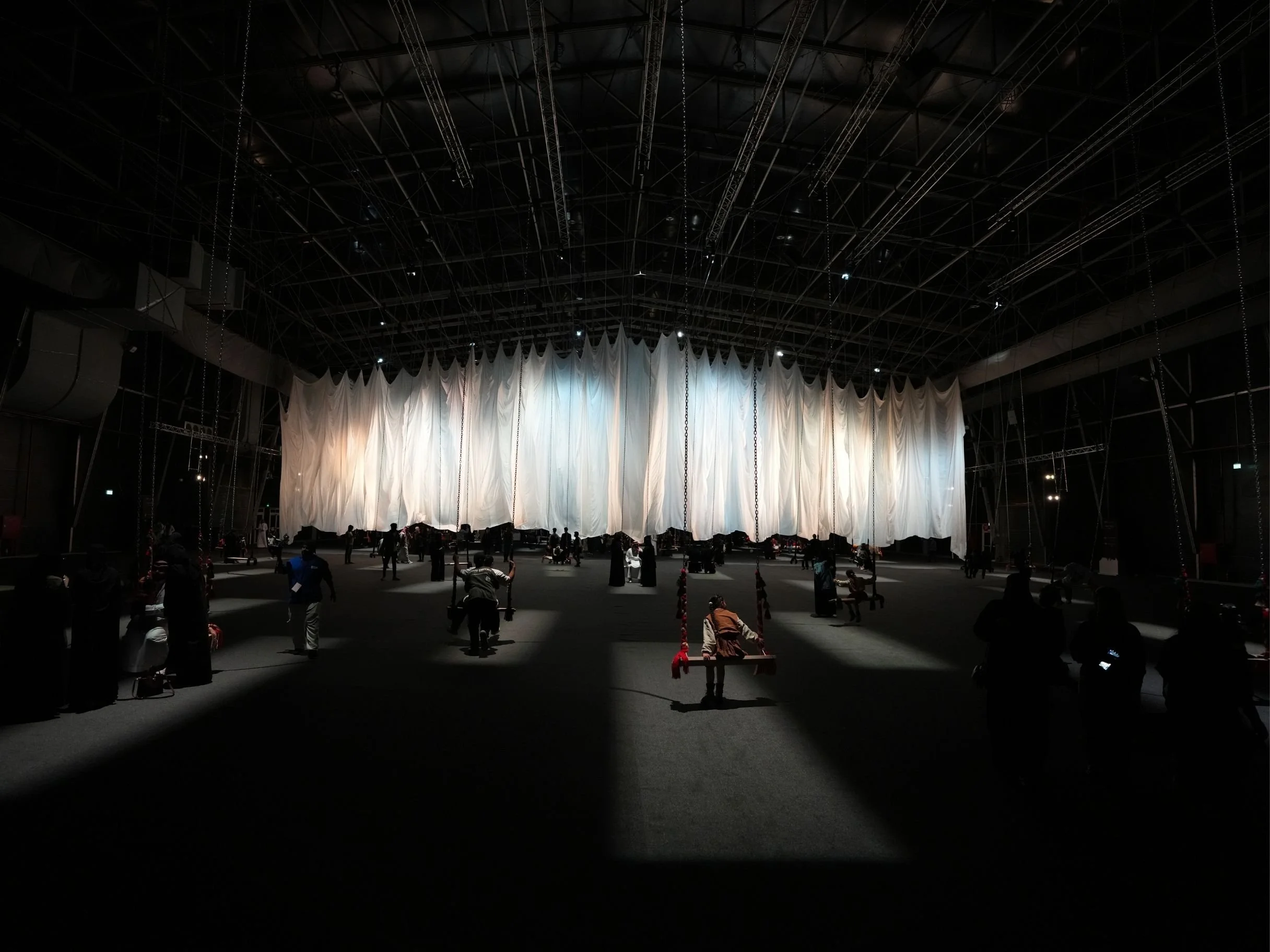 Large stage with white curtains and suspension cables in a dark auditorium, with a few people around.