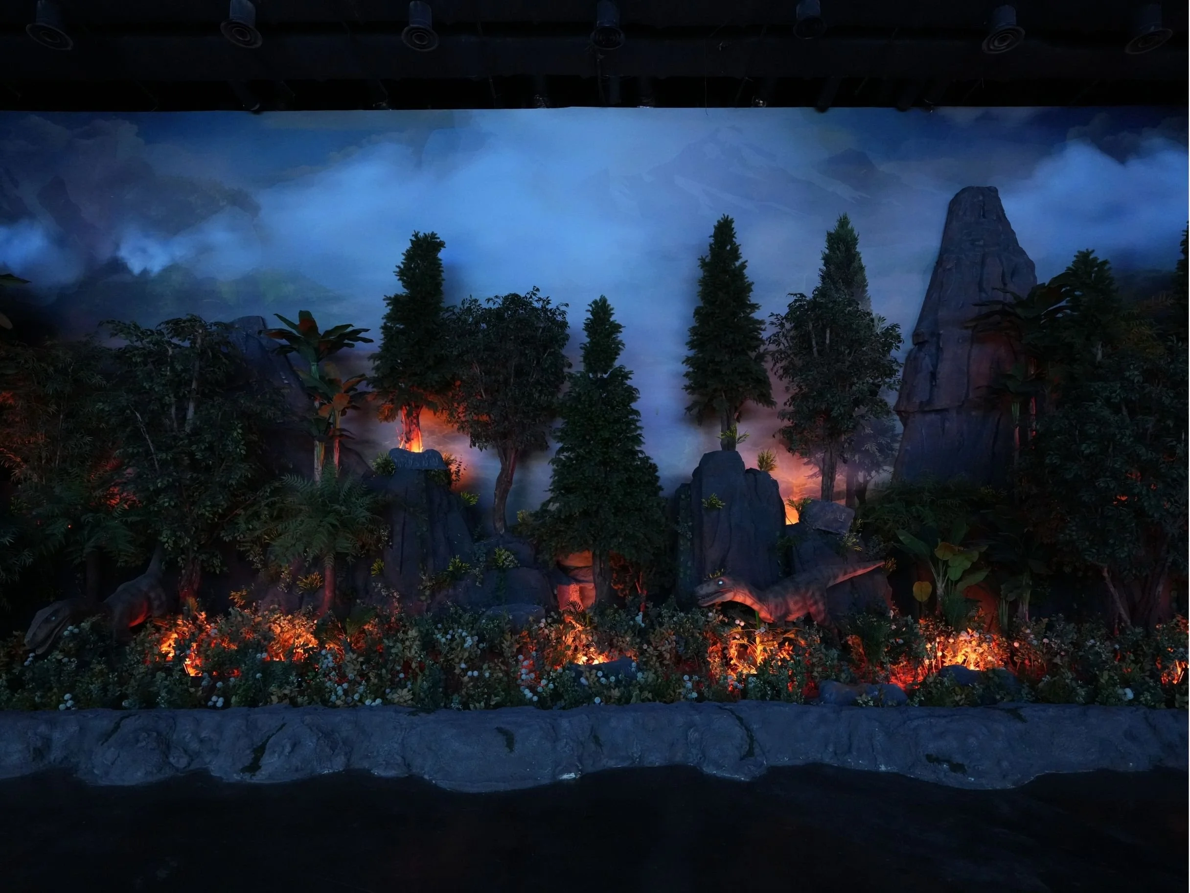 Indoor setting with a large scenic nature mural of mountains, trees, and fog, illuminated by colorful lighting at the bottom, likely part of an exhibit or themed space.