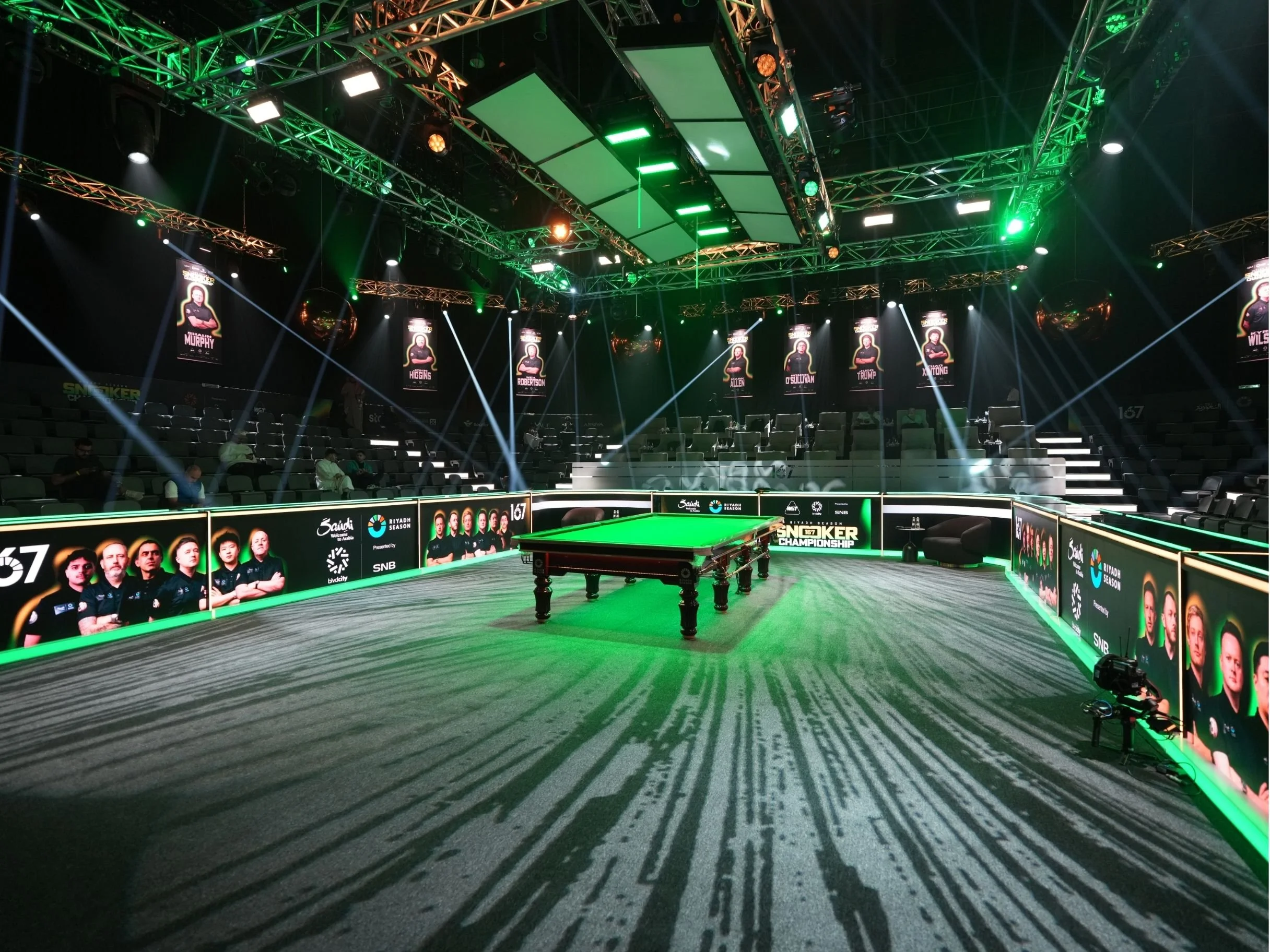  snooker, boulevard world, sela, sauri arabia, riyadh, surrounded by seating and multiple large screens displaying players and event information. The arena is decorated with green lighting, banners, and posters for a professional e-sports tournament