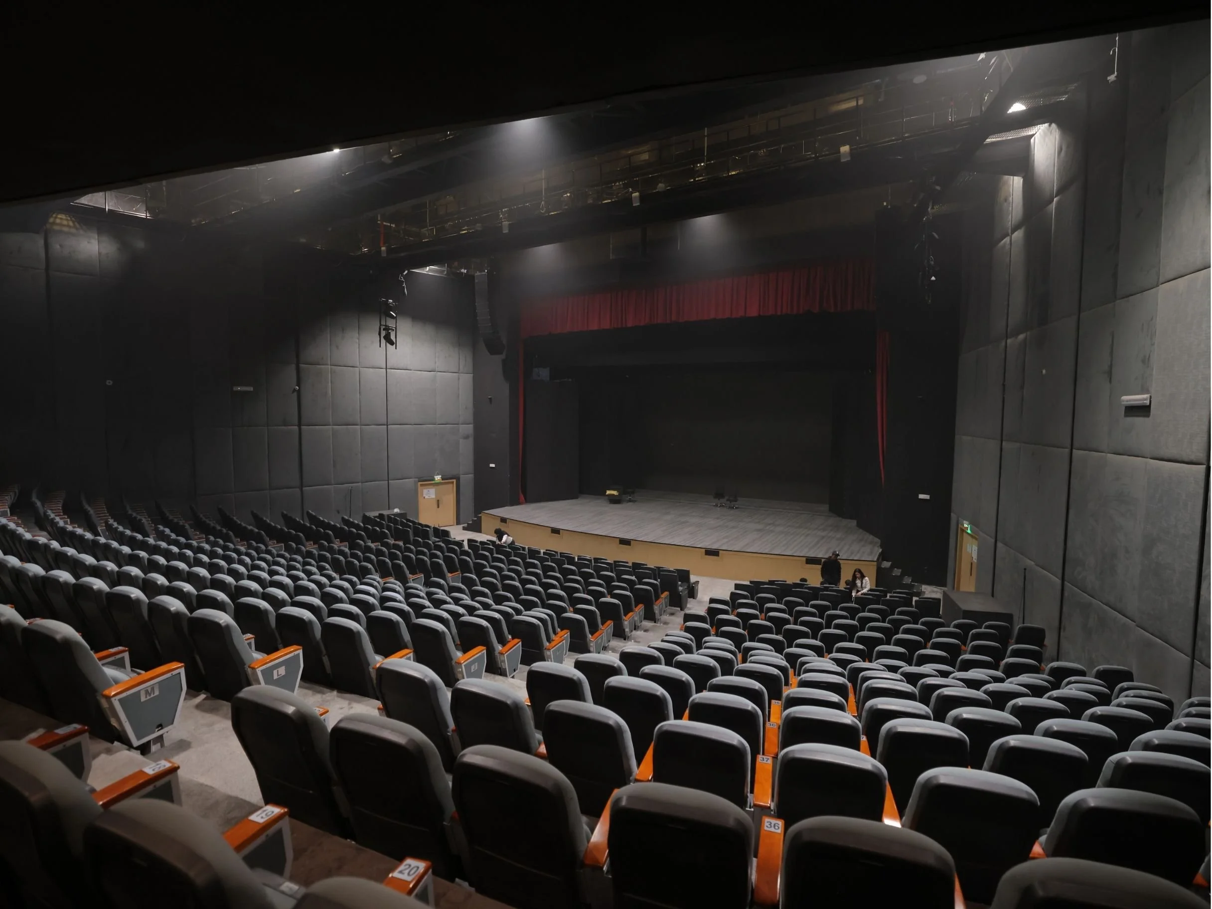 Empty theater auditorium with rows of black seats, a stage with red curtains, and overhead lighting in a dimly lit room. bakr al sheddi, bouvard city