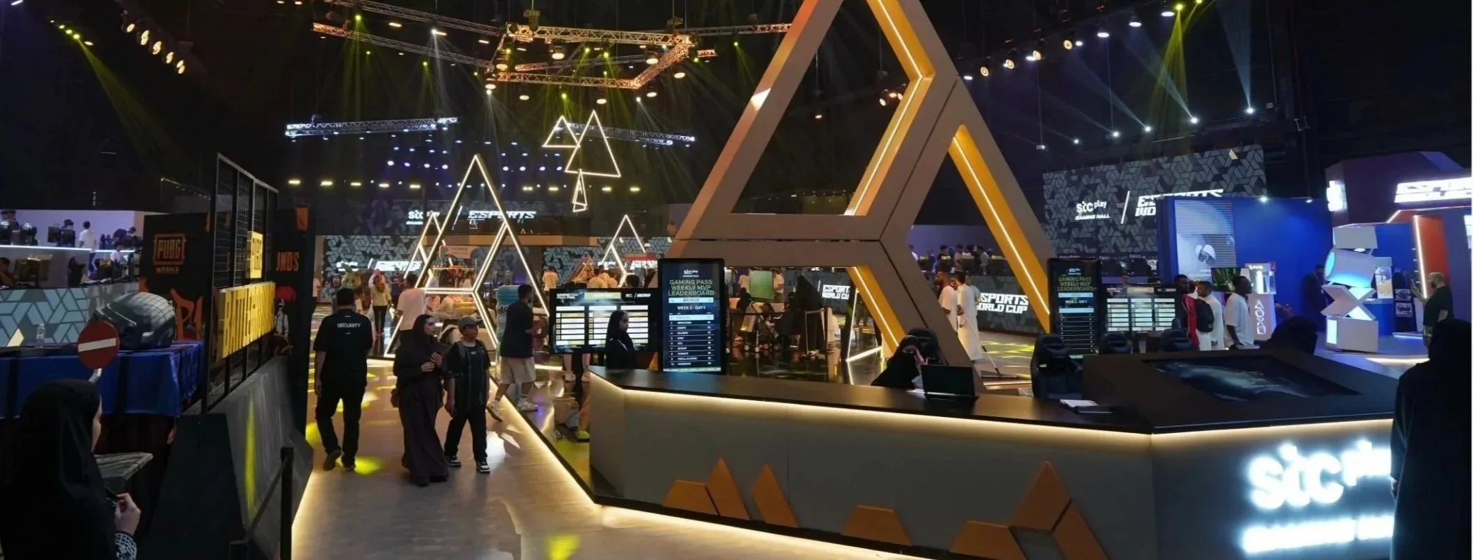An indoor gaming or tech event with illuminated geometric decorations and signage for e-sports or gaming brands, with people walking and standing around. stc gaming lounge. EWC, saudi arabia