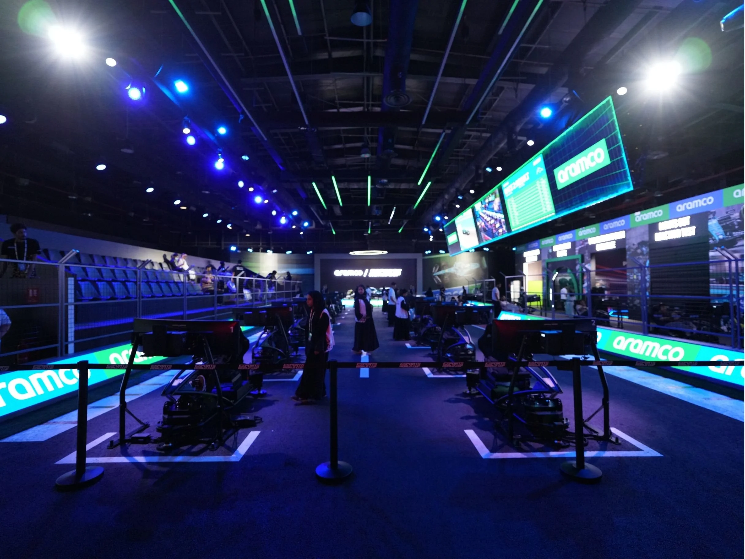 Indoor esports arena with multiple screens displaying 'Aramco' logo, gaming stations with seats, and blue and green stage lighting. aramco, boulevard world, event, set design, construction