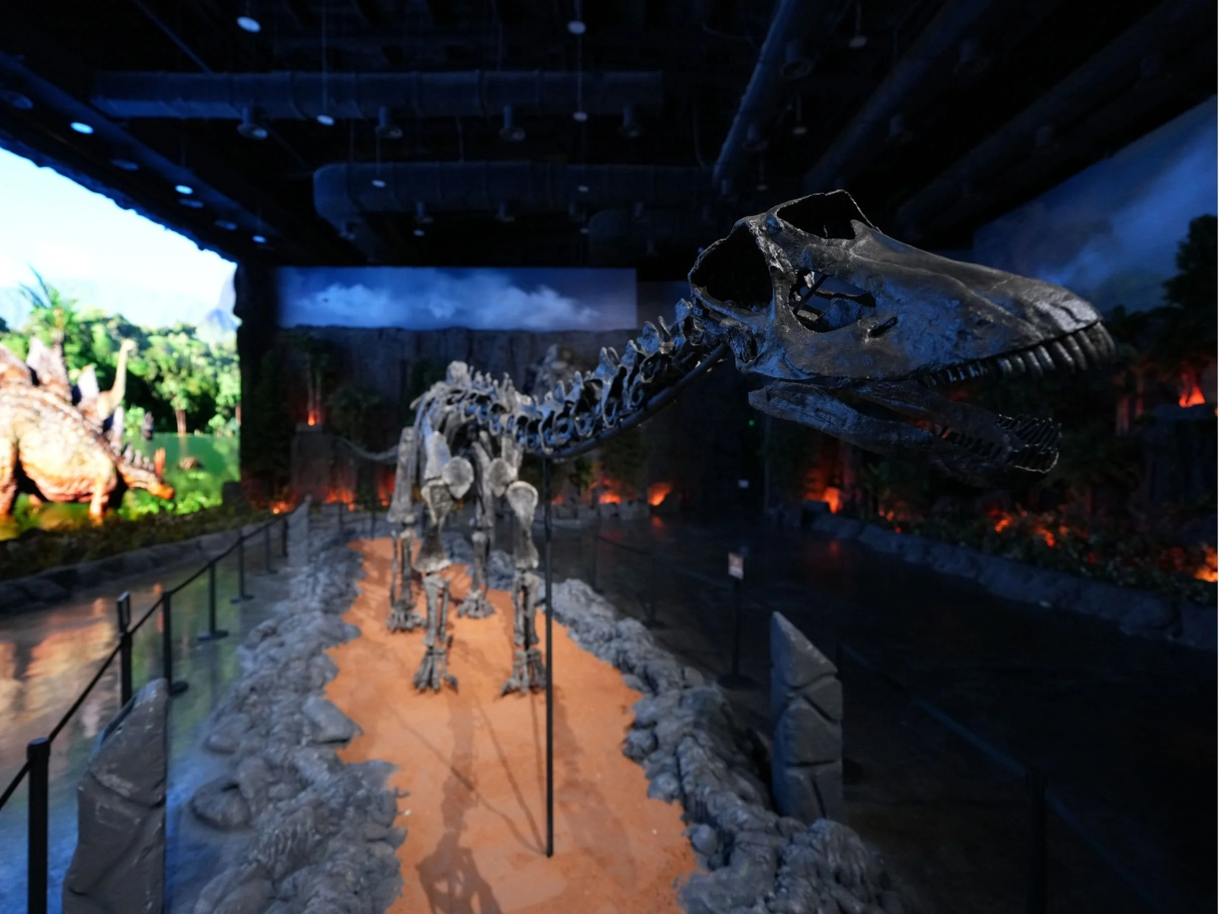 A dinosaur skeleton, likely a theropod, displayed in a museum with a dark ceiling, spotlighting the bones, and a background with a digital landscape scene.