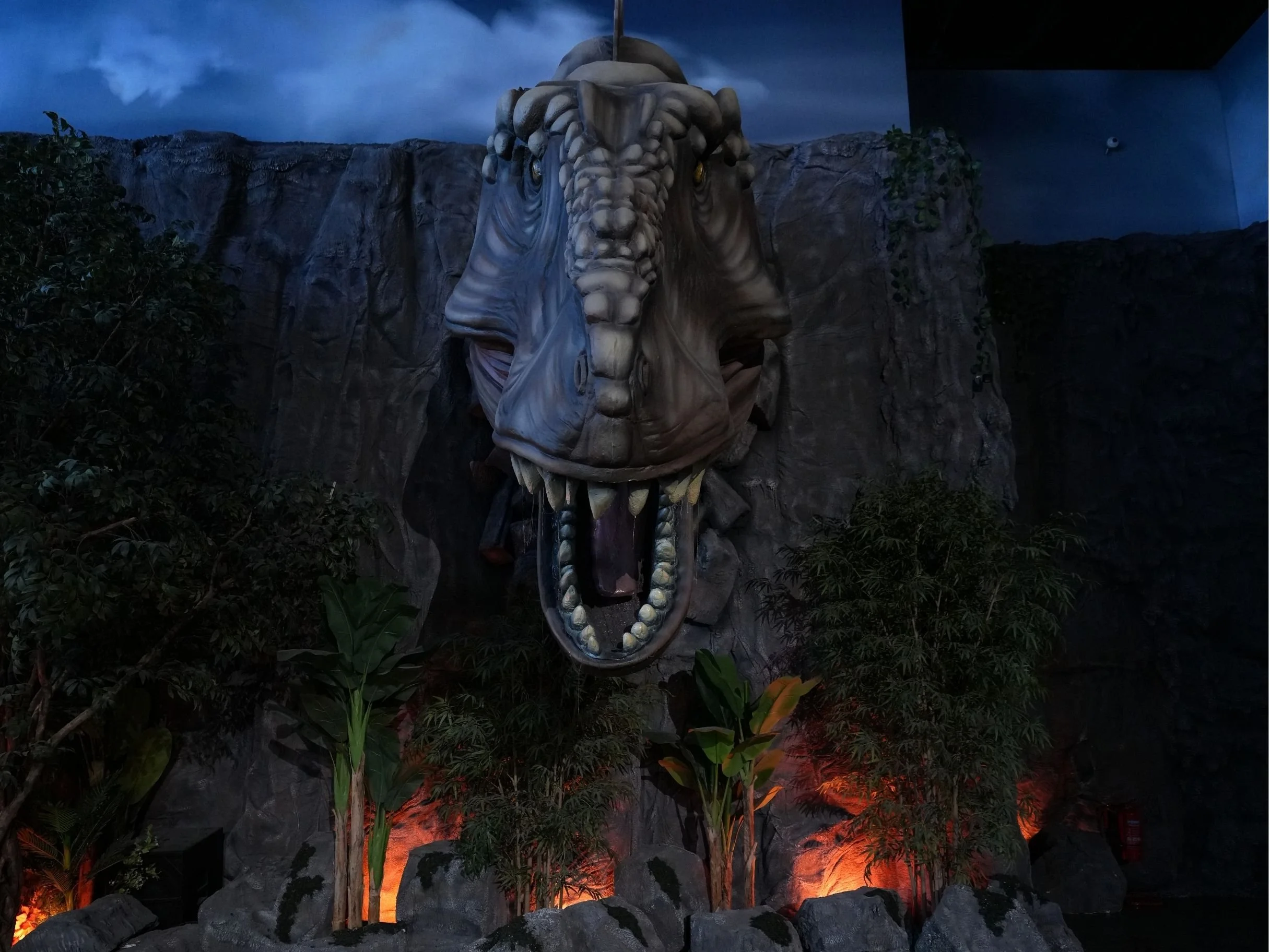 A large tiger head sculpture with an open mouth showing teeth, set against a rocky background and surrounded by jungle plants, illuminated by colorful lights.