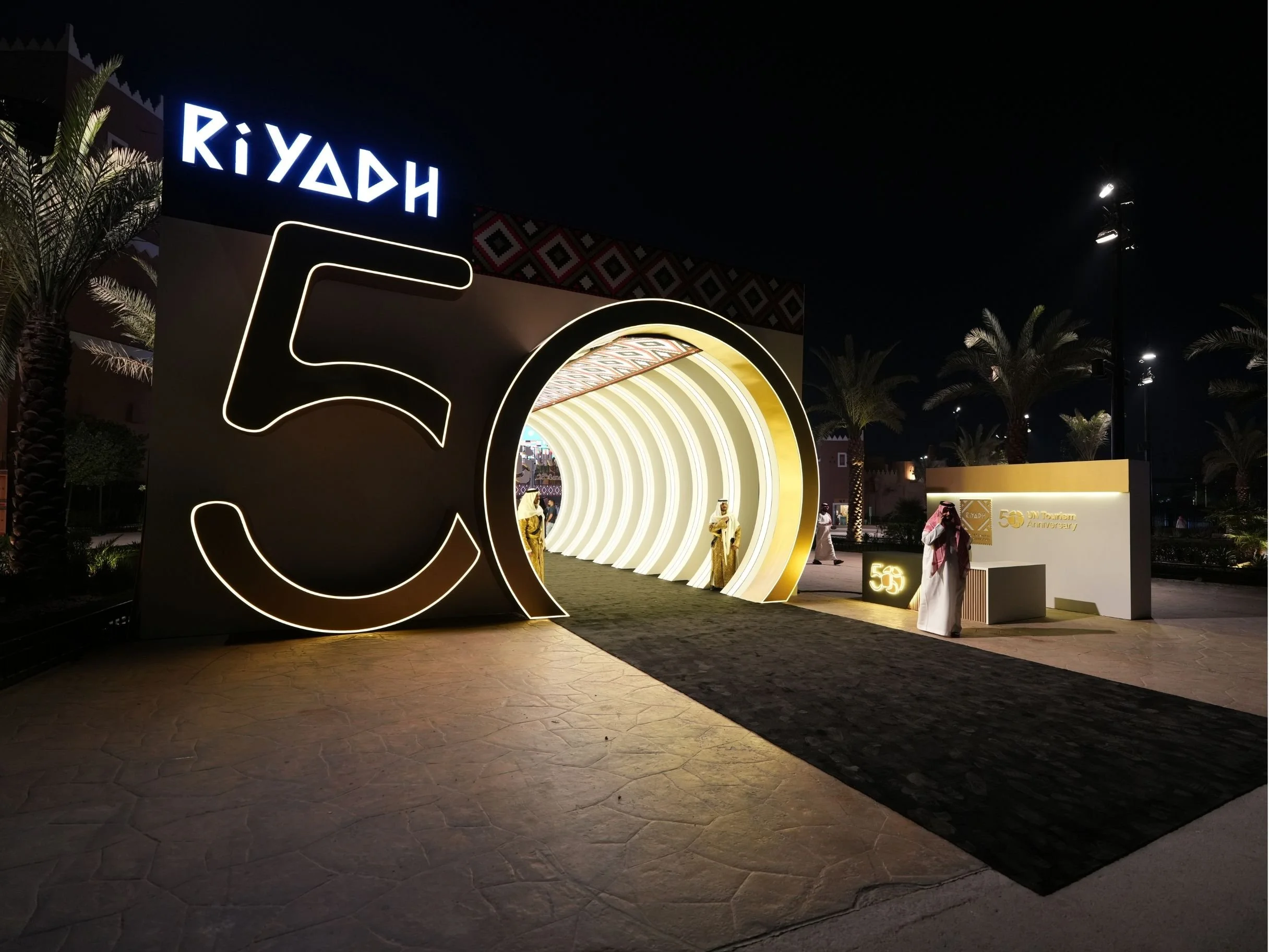 Nighttime view of a decorated entrance with illuminated neon signs celebrating 50 years, palm trees, and a dark sky.boulevard world, sela, construction