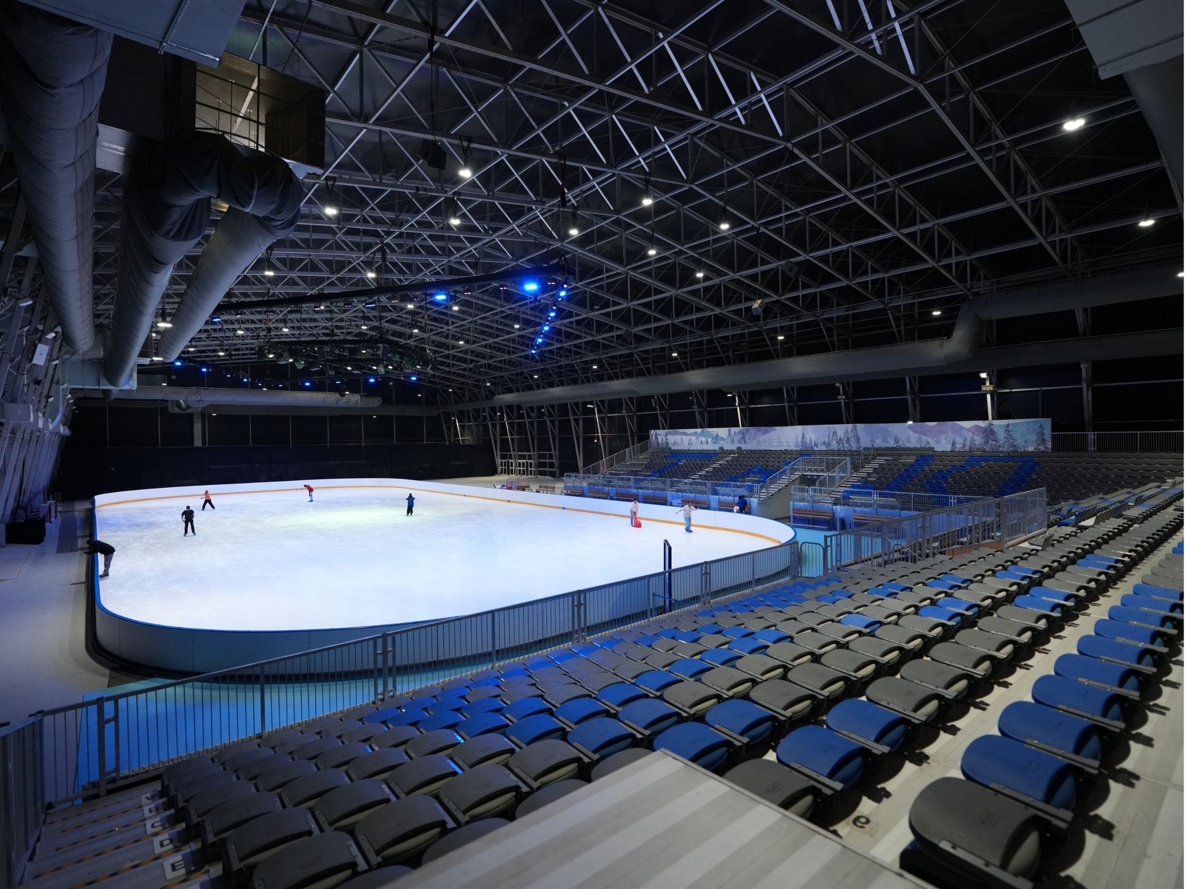 An indoor ice skating rink with a few skaters on the ice. The arena is enclosed with a high, metal lattice ceiling and blue lighting. Empty blue and gray seats are visible around the rink. cool arena iceskating rink.