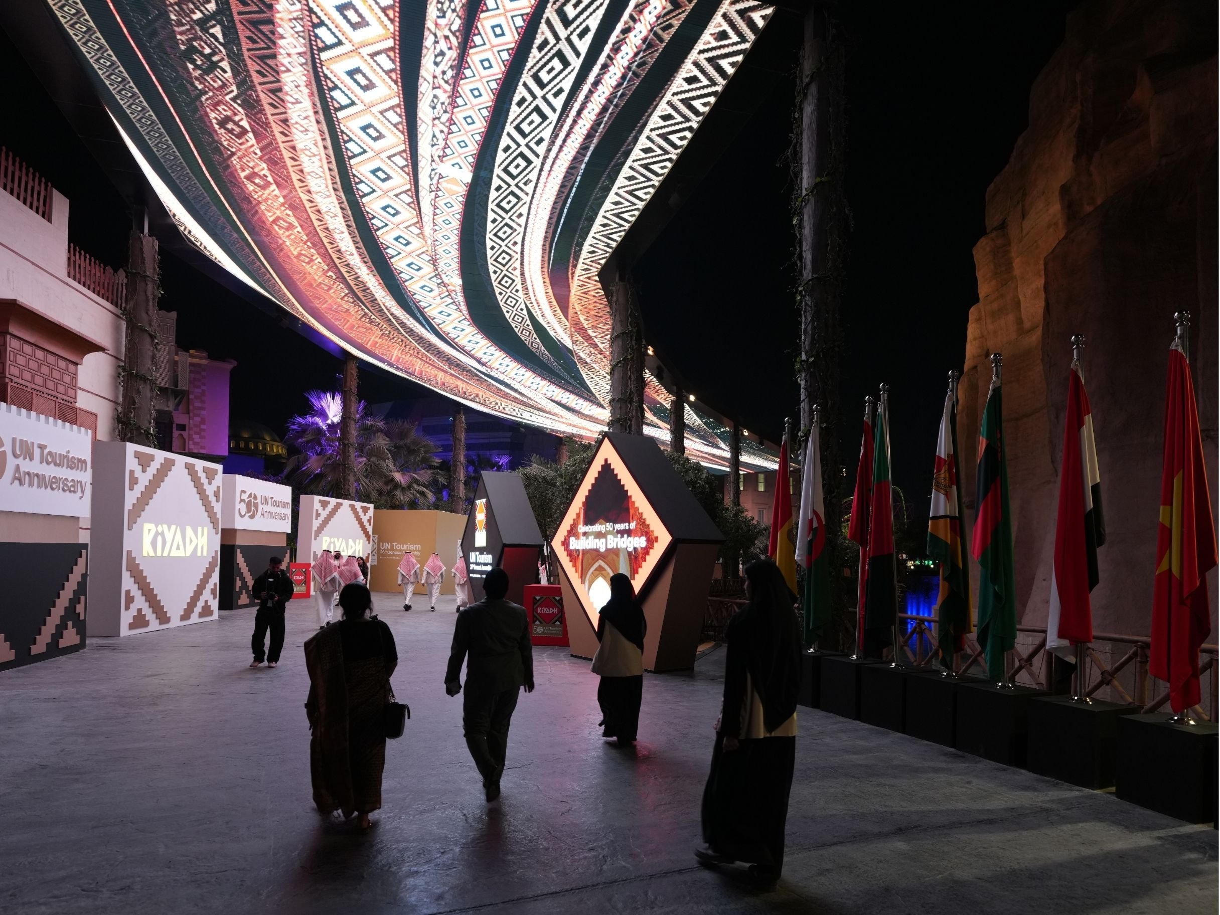 A nighttime outdoor event with people walking under a large decorative canopy with colorful geometric patterns. There are different flags on display, and illuminated signs celebrating 50 years of building bridges and a UN Tourism anniversary.