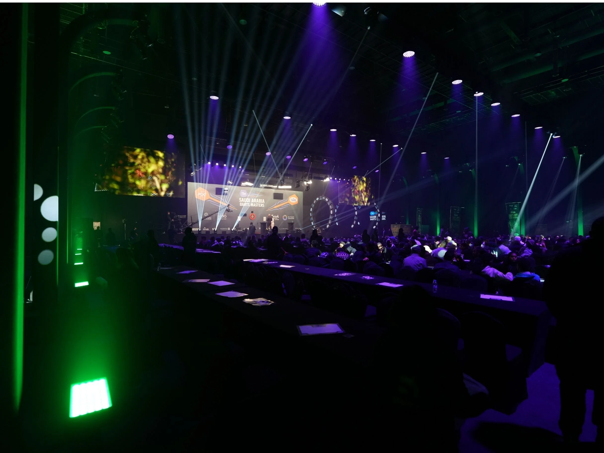 A stage with colorful lights and large screens at the Saudi Arabia Darts Masters event, and an audience seated in front.