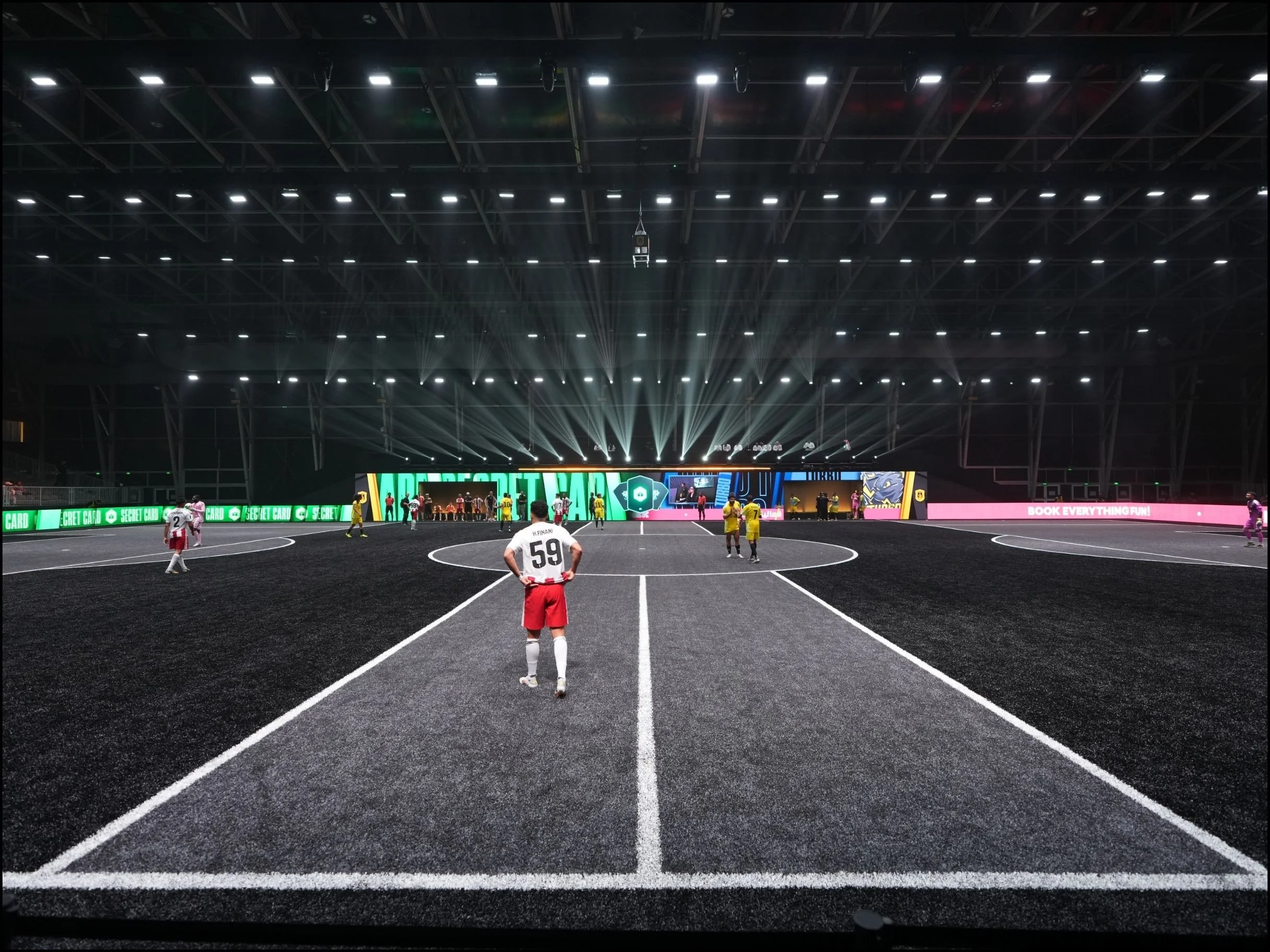 Indoor sports arena with a black synthetic running track, players on the field, LED advertising screens along the sides, bright overhead lighting, and a scoreboard. light design, set design, event, kings league.