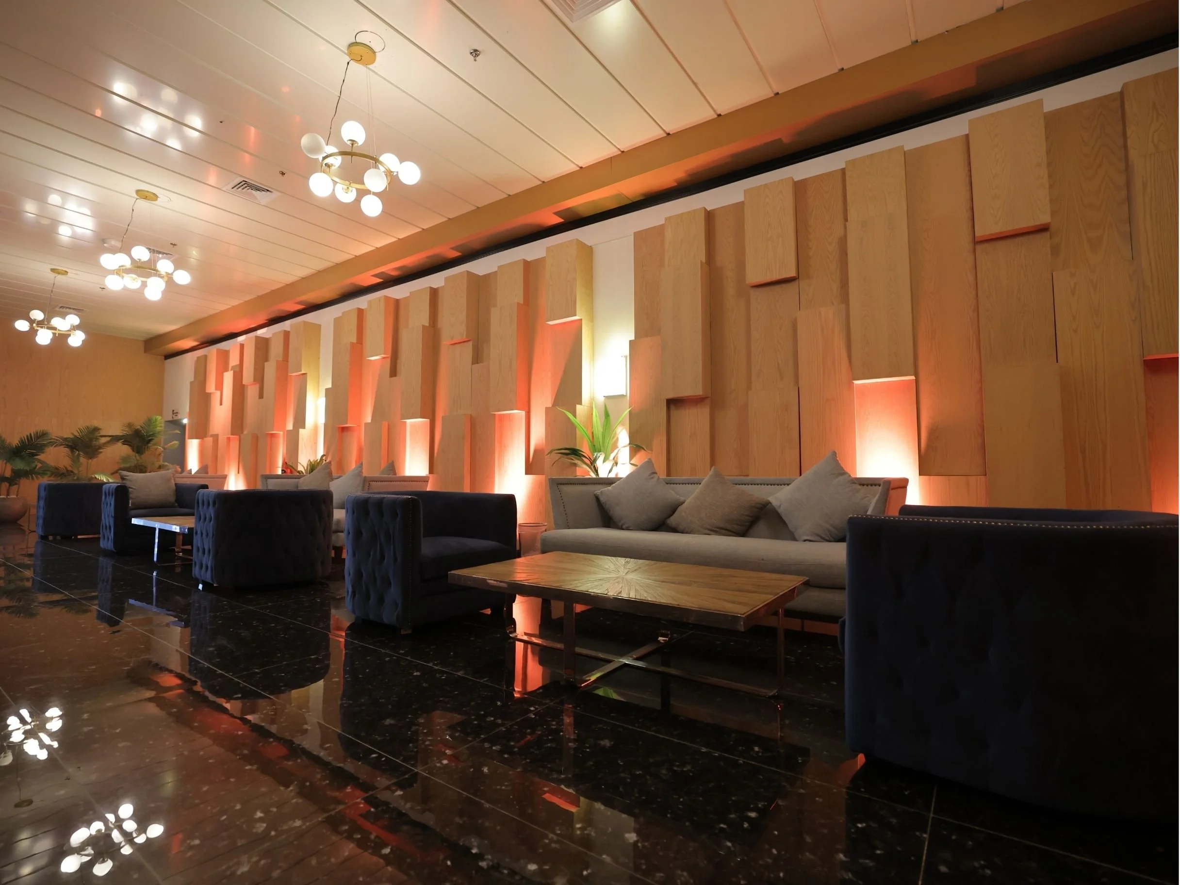 Interior of a modern lounge or lobby area with black marble flooring, wooden wall panels, plush sofas, and decorative plants. Multiple ceiling lights and a chandellier provide lighting.bakr al sheddi, bouvard city