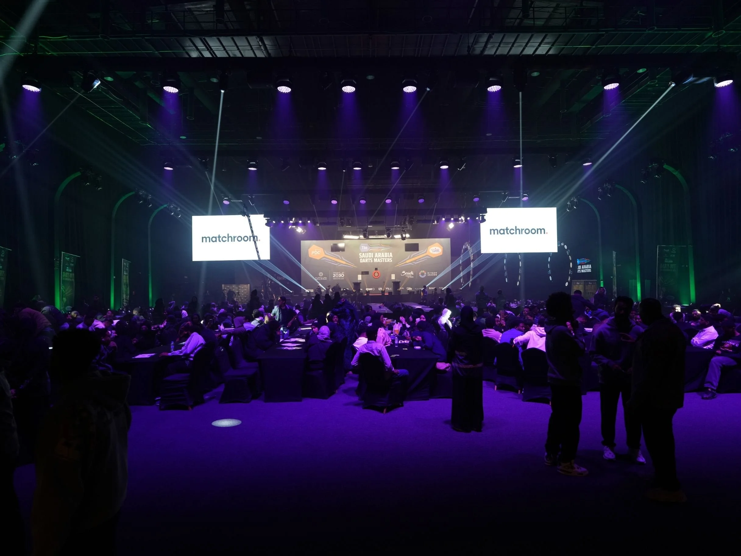 An indoor stage with purple and green lighting, two large screens displaying 'matchroom', and an audience sitting at tables in a darkened room.