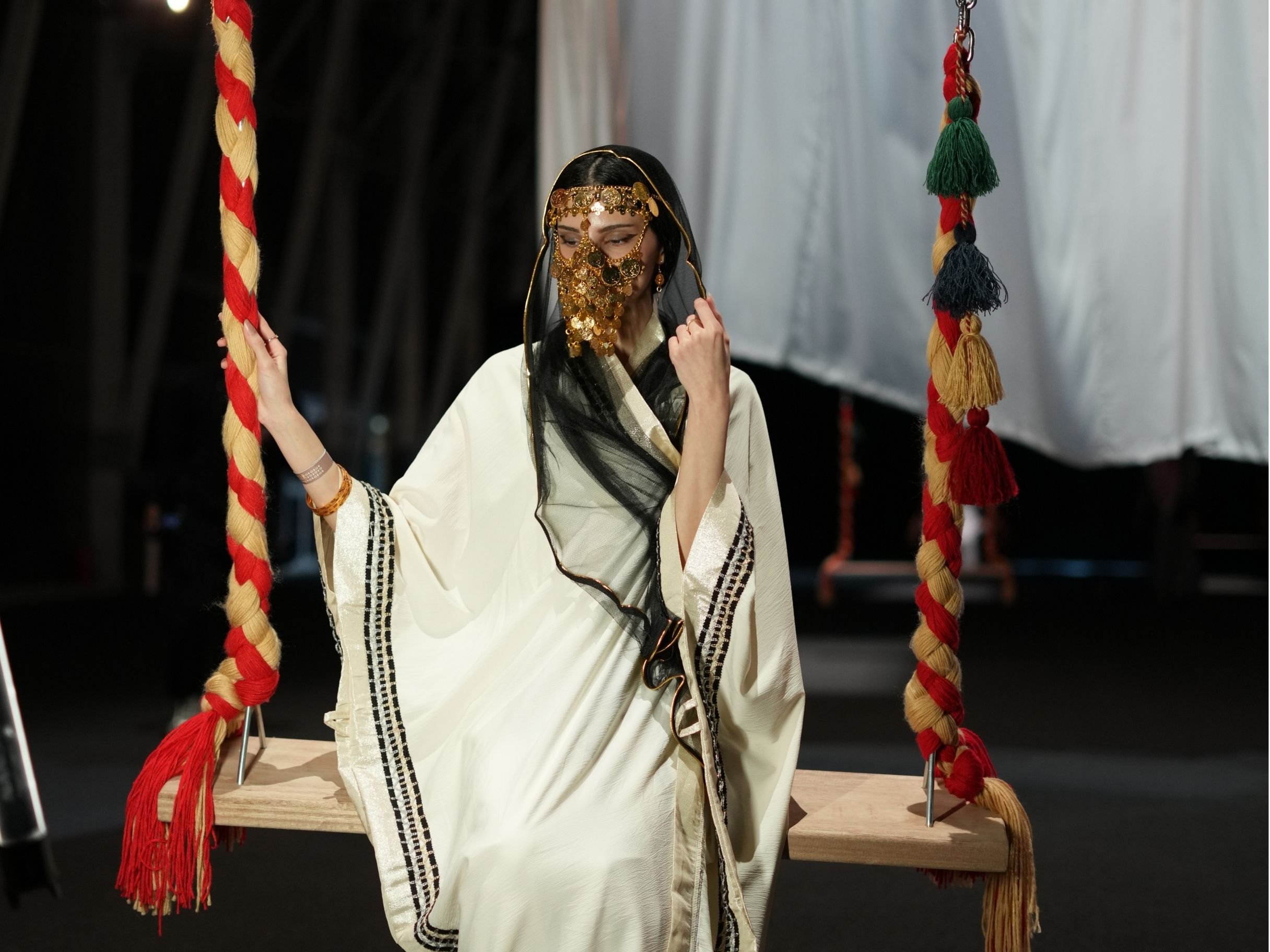 A woman dressed in a traditional costume with gold jewelry, sitting on a swing decorated with red and yellow braided ropes, against a dark background.