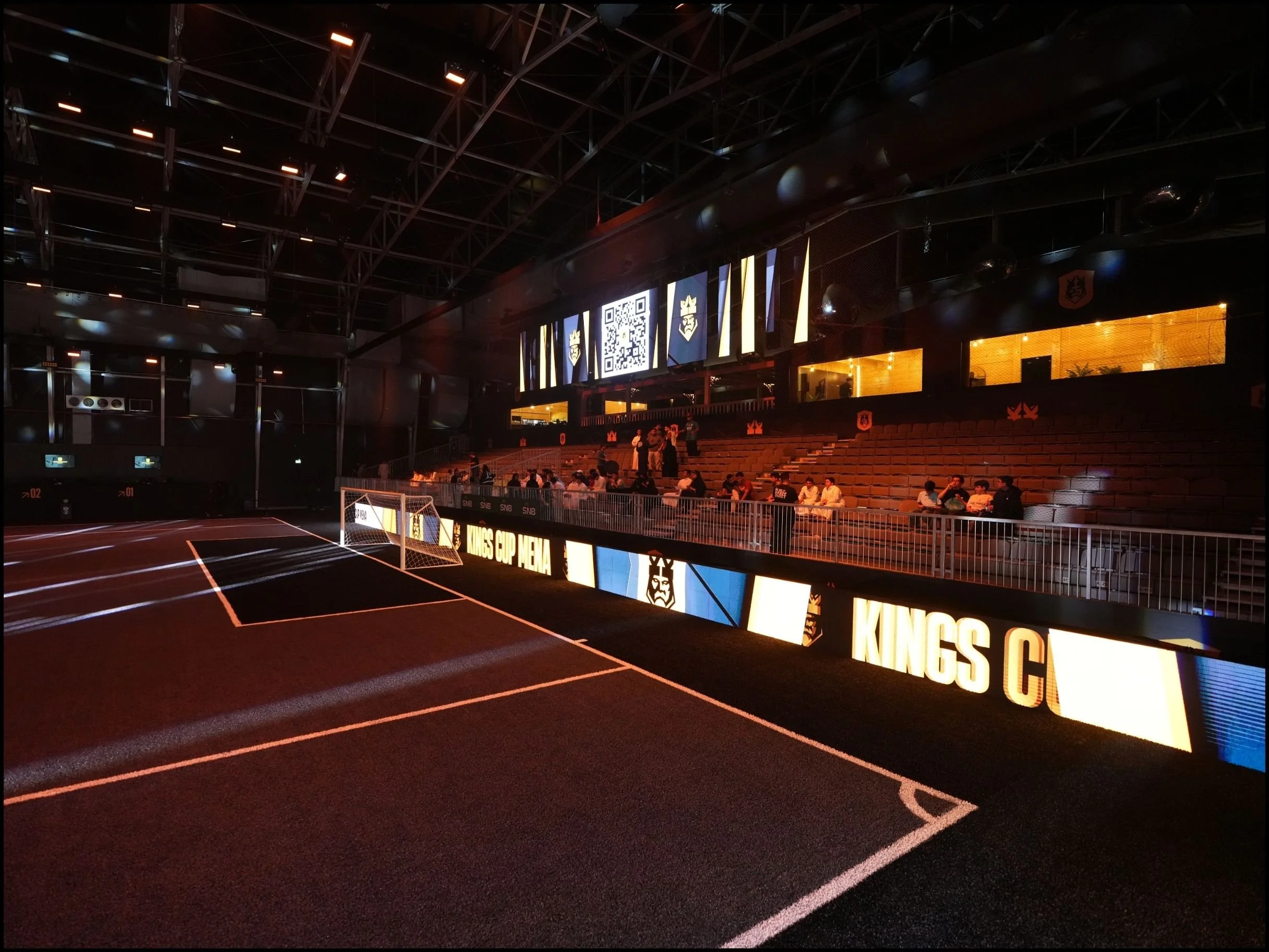 Indoor sports arena with a small soccer goal, illuminated LED screens displaying logos and a QR code, and tiered seating with some spectators, under a metallic ceiling structure.