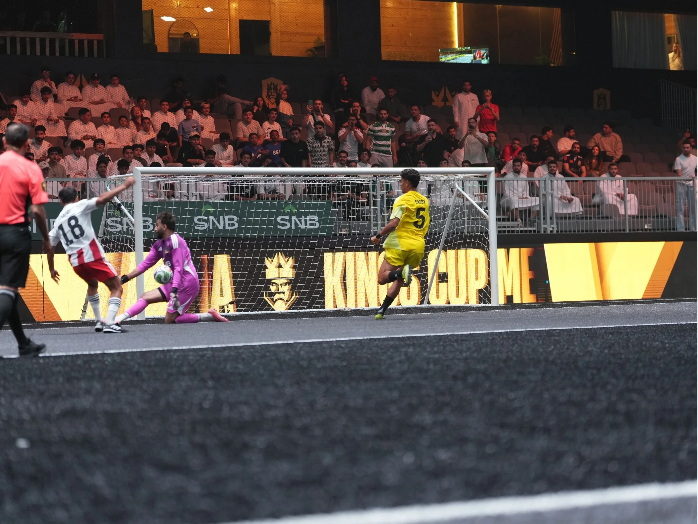 A soccer game in progress with players near the goal, spectators watching from the stands, and a referee overseeing the match. The scene takes place inside a stadium with advertisements and a digital display in the background.