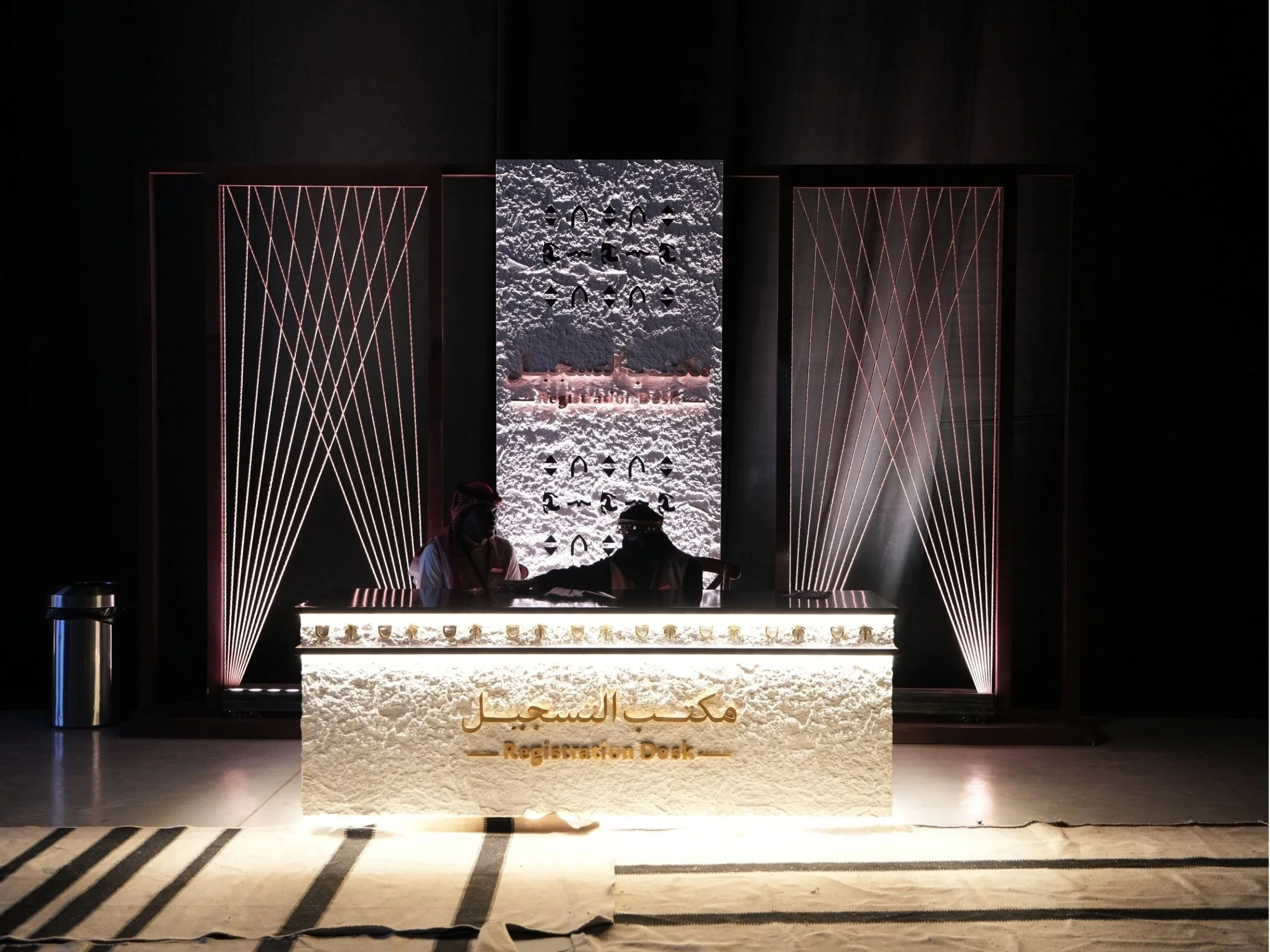 A reception desk in a dark room with decorative lighting and a sign that reads "Registration Desk" in Arabic and English, with two people sitting behind the desk.