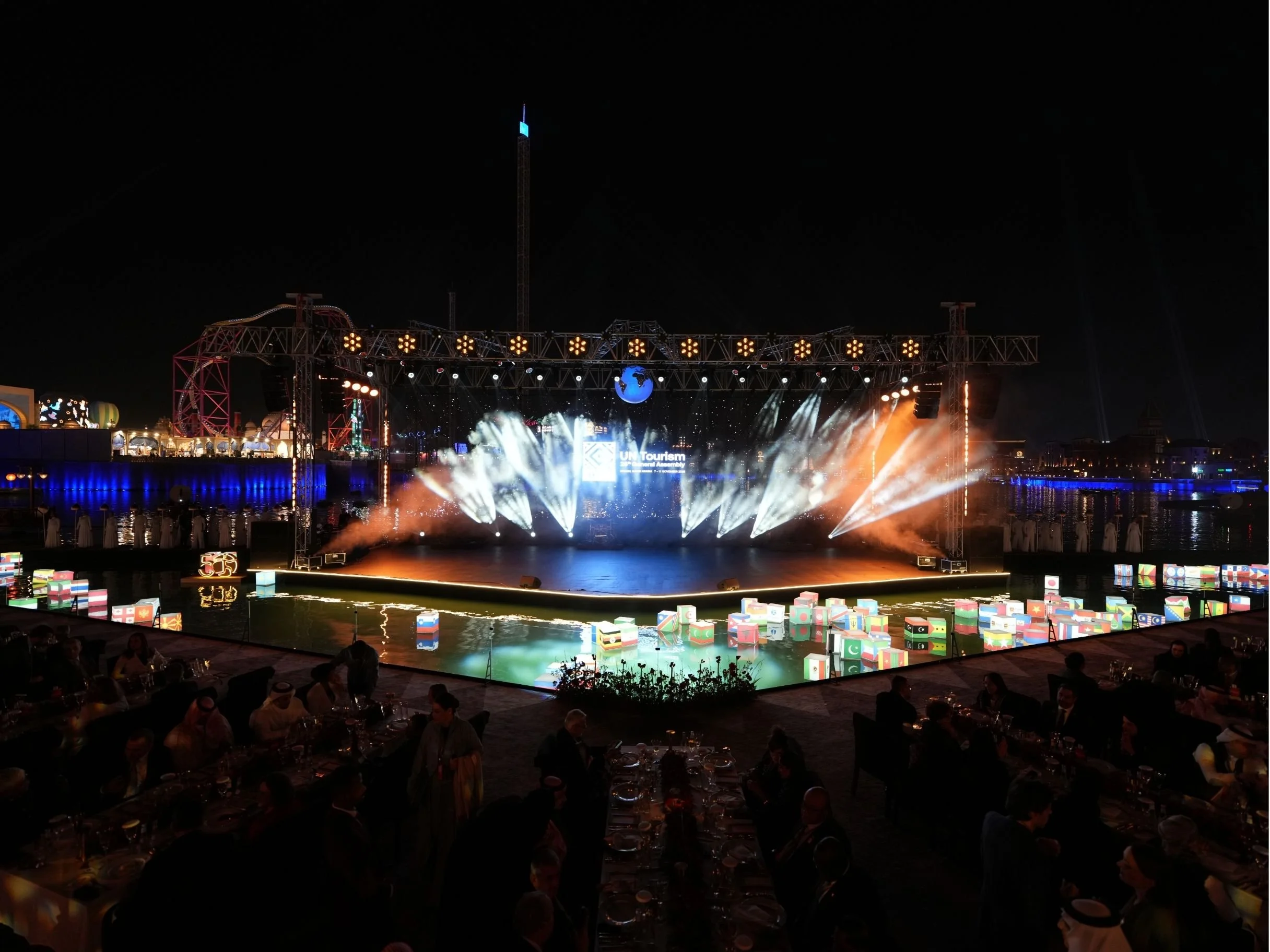 Nighttime outdoor event with a stage, colorful lights, fog effects, an astronaut figure, and international flags on floating cubes, with seated audience in front. boulevard world, un tourism, 5oth anniversary.