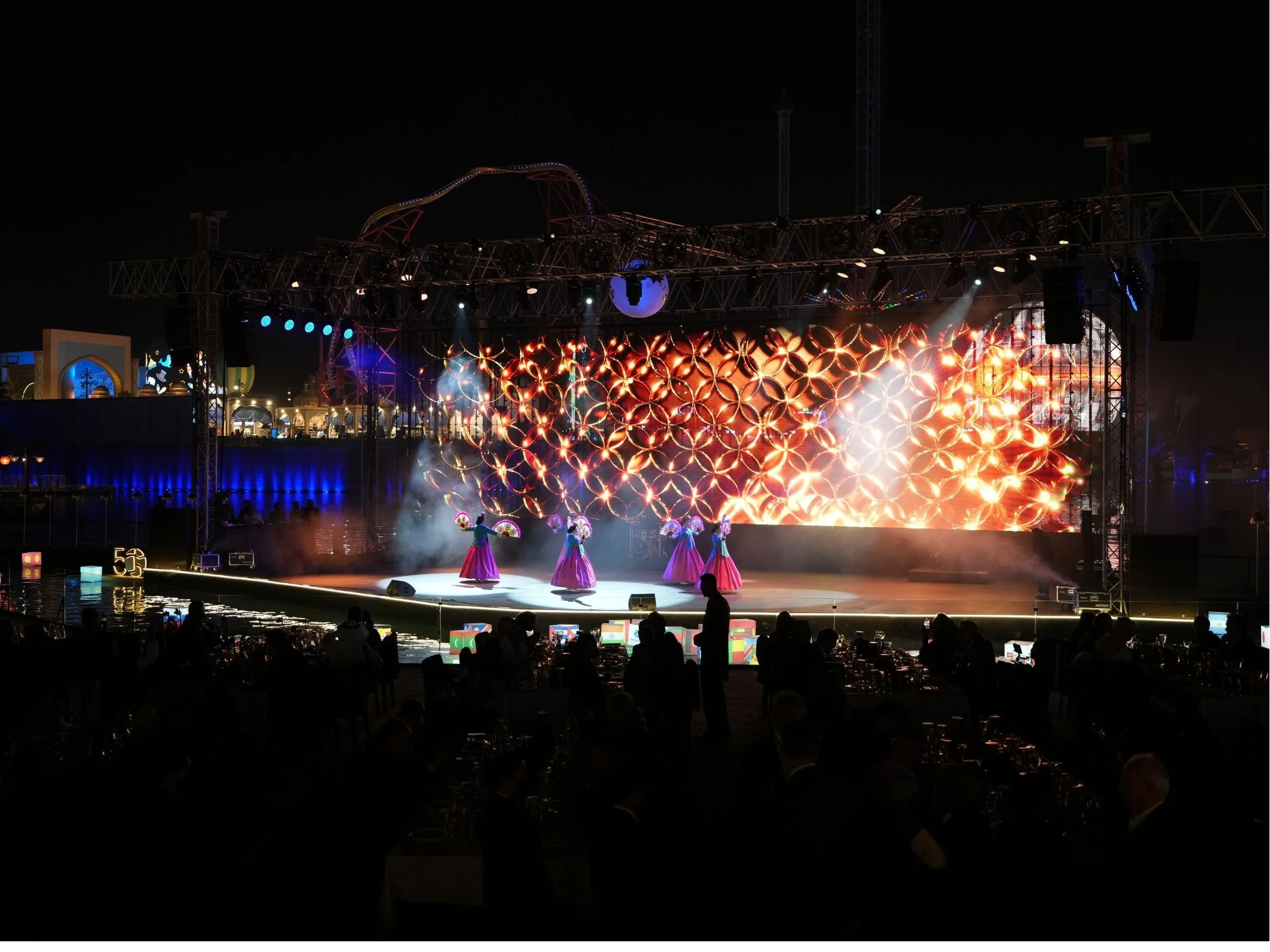 Nighttime stage performance with dancers in traditional costumes, large digital screen displaying bright orange and red patterns, boulevard world, sela, construction