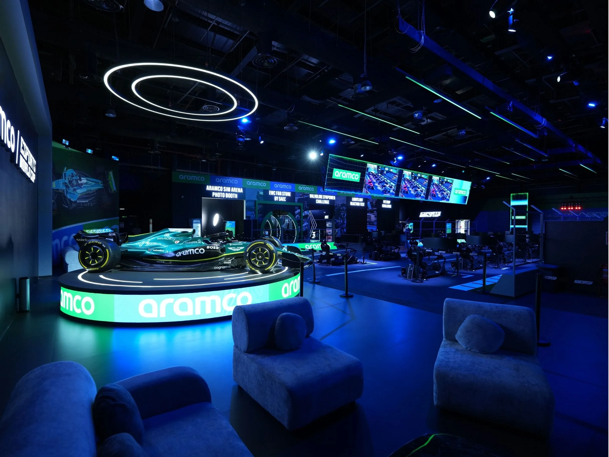 A dark indoor event space with a race car on a platform with bright green accents, branded with 'Aramco,' surrounded by plush chairs and illuminated by futuristic circular and linear LED lights.  aramco, boulevard world, event, set design, constructi
