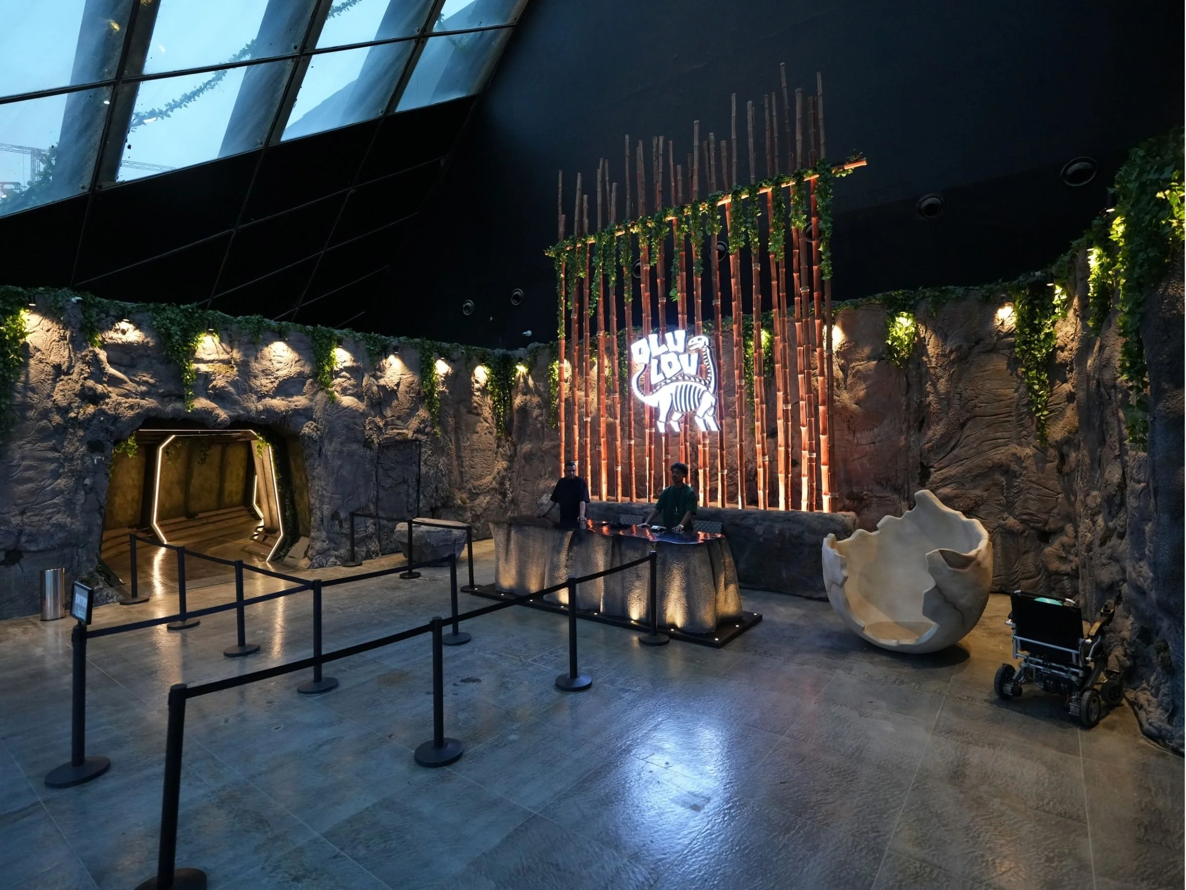 Indoor scene with a rocky wall, illuminated signage featuring a dinosaur illustration, and greenery. There are people with a stroller and a barricade separating the space from visitors.