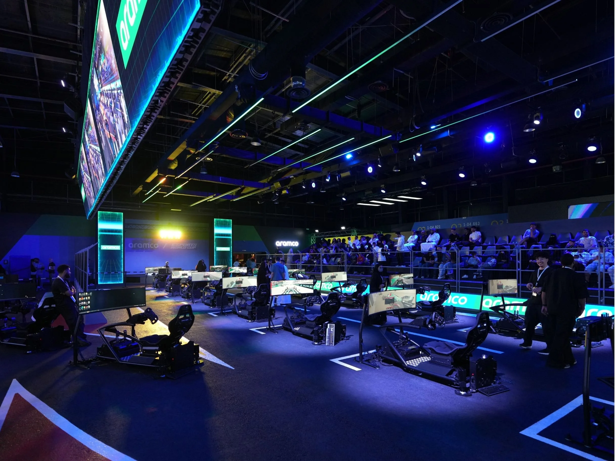 An indoor gaming or esports arena with racing game simulators and seating area illuminated by blue and green LED lights. aramco, boulevard world, event, set design, construction