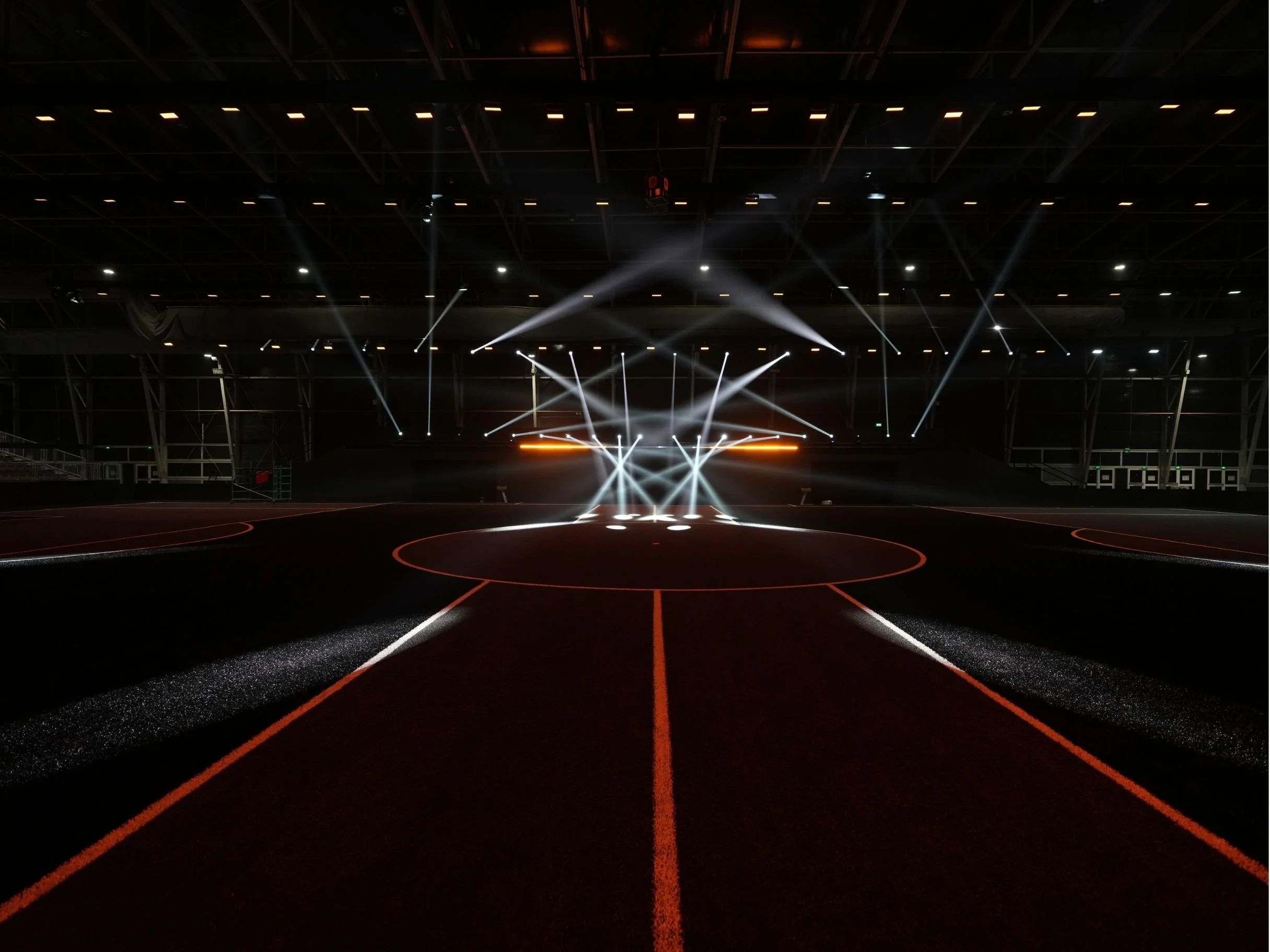An empty sports arena with track markings, bright overhead lighting, and a stage with multiple spotlights and equipment for a concert or event.