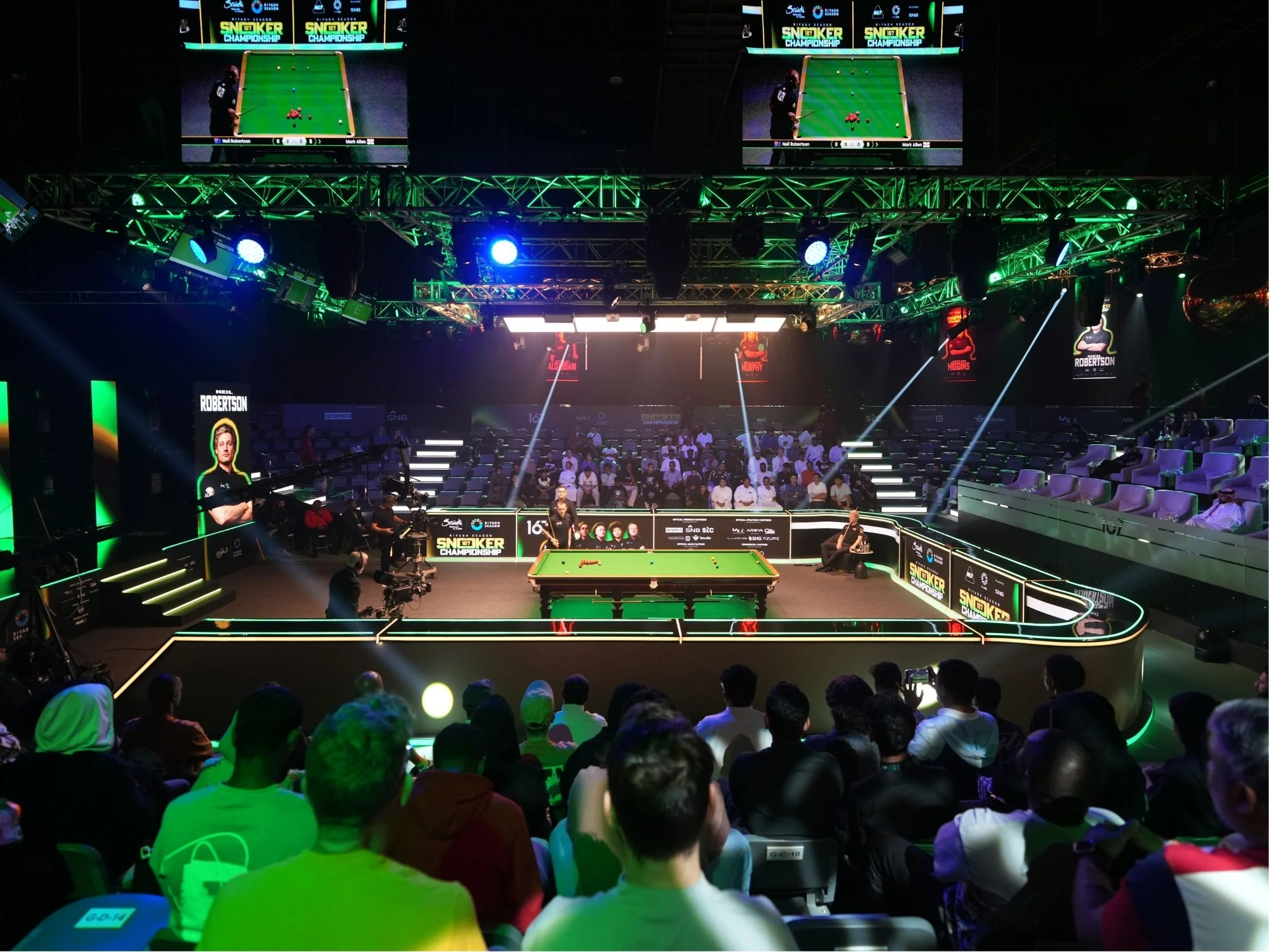 An indoor snooker tournament with a large audience, multiple screens displaying the game, and players competing on a snooker table under vibrant lighting. snookers, riyadh, saudi arabia, 
