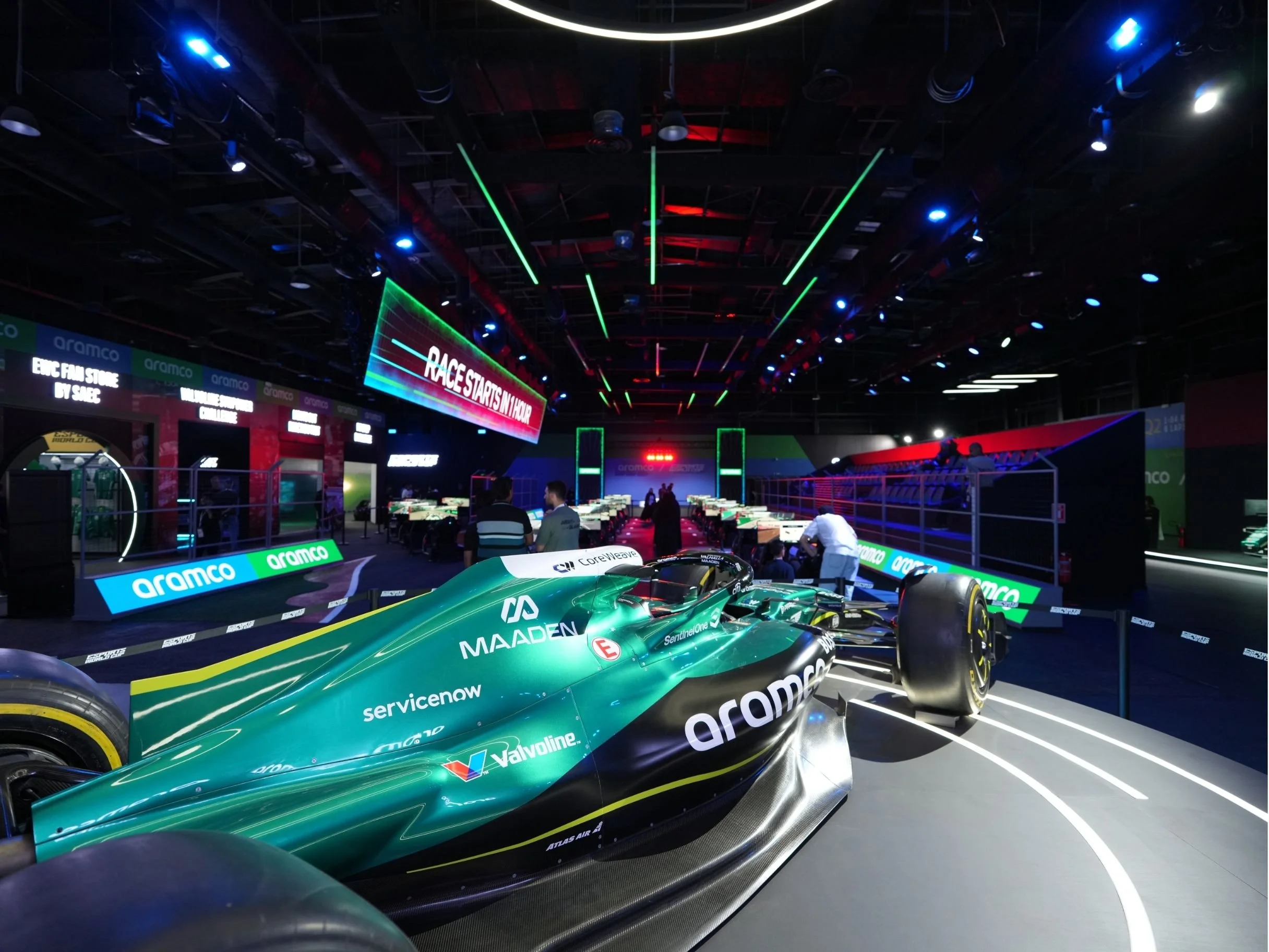 Indoor racing event with a racing car in the foreground, digital screens displaying race information, vibrant lighting, and spectators in the background. aramco, boulevard world, event, set design, construction